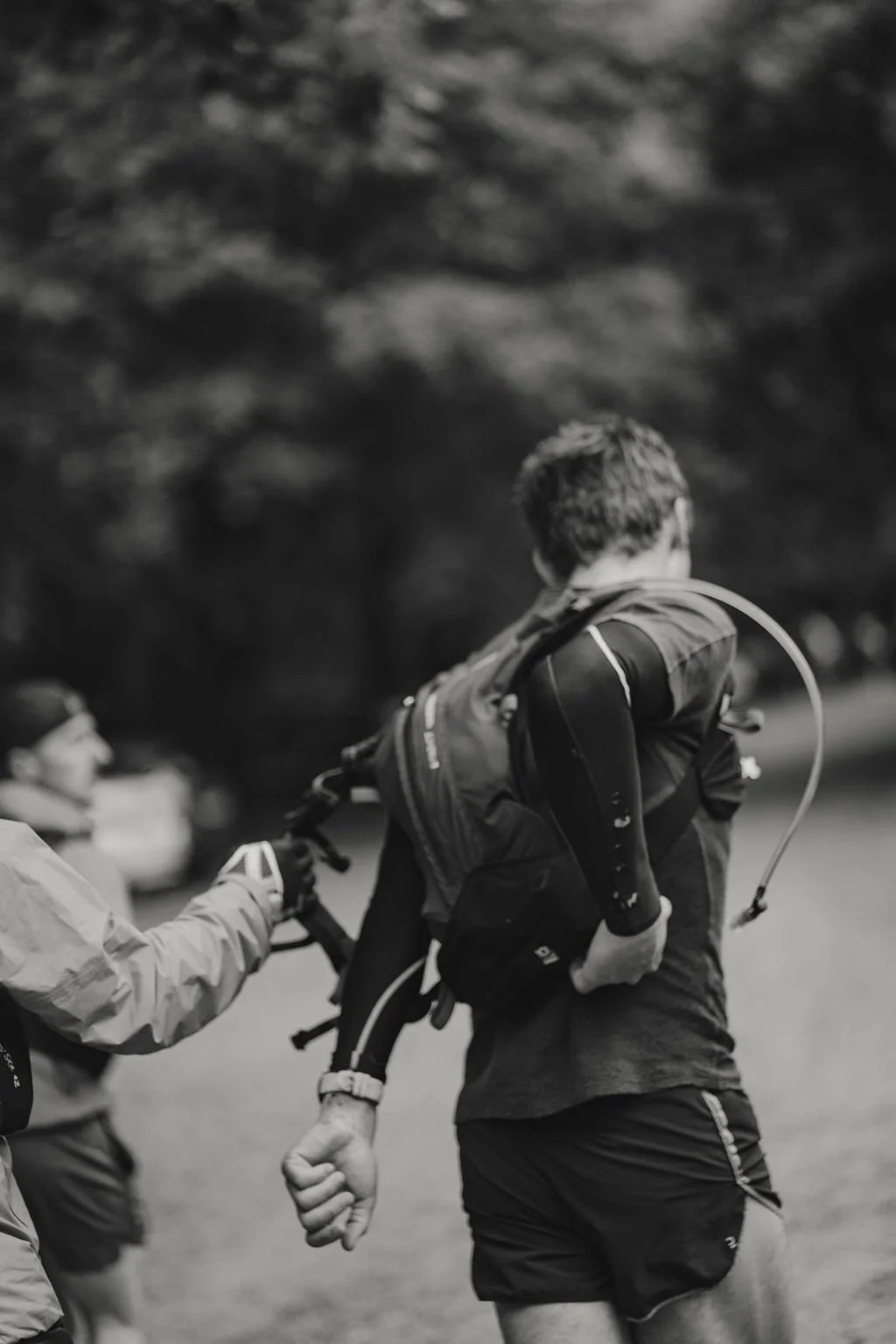 A person running outdoors with a backpack, wearing athletic clothes, during a trail or race, with trees and other runners in the background.