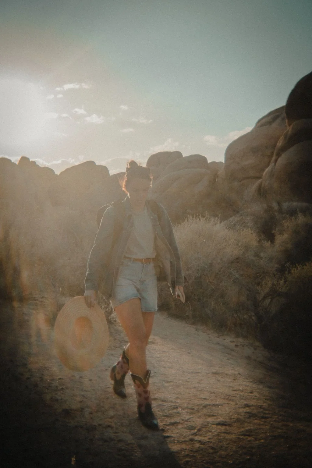 A person walking along a dirt trail in a desert landscape with rocks and dry bushes, holding a wide-brimmed straw hat and wearing cowboy boots, with the sun low in the sky.