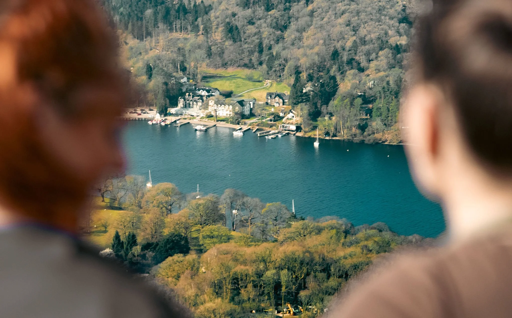 Two people with blurred faces looking at a scenic view of a lake, boats, trees, and houses on the opposite shore.