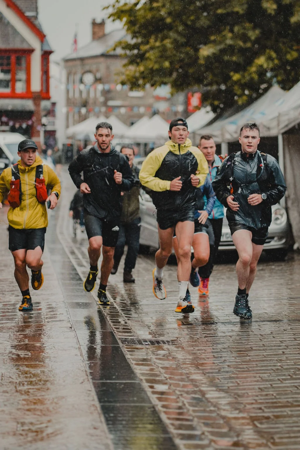 Group of runners participating in a race on a rainy day in an urban area with wet cobblestone streets and tents in the background.