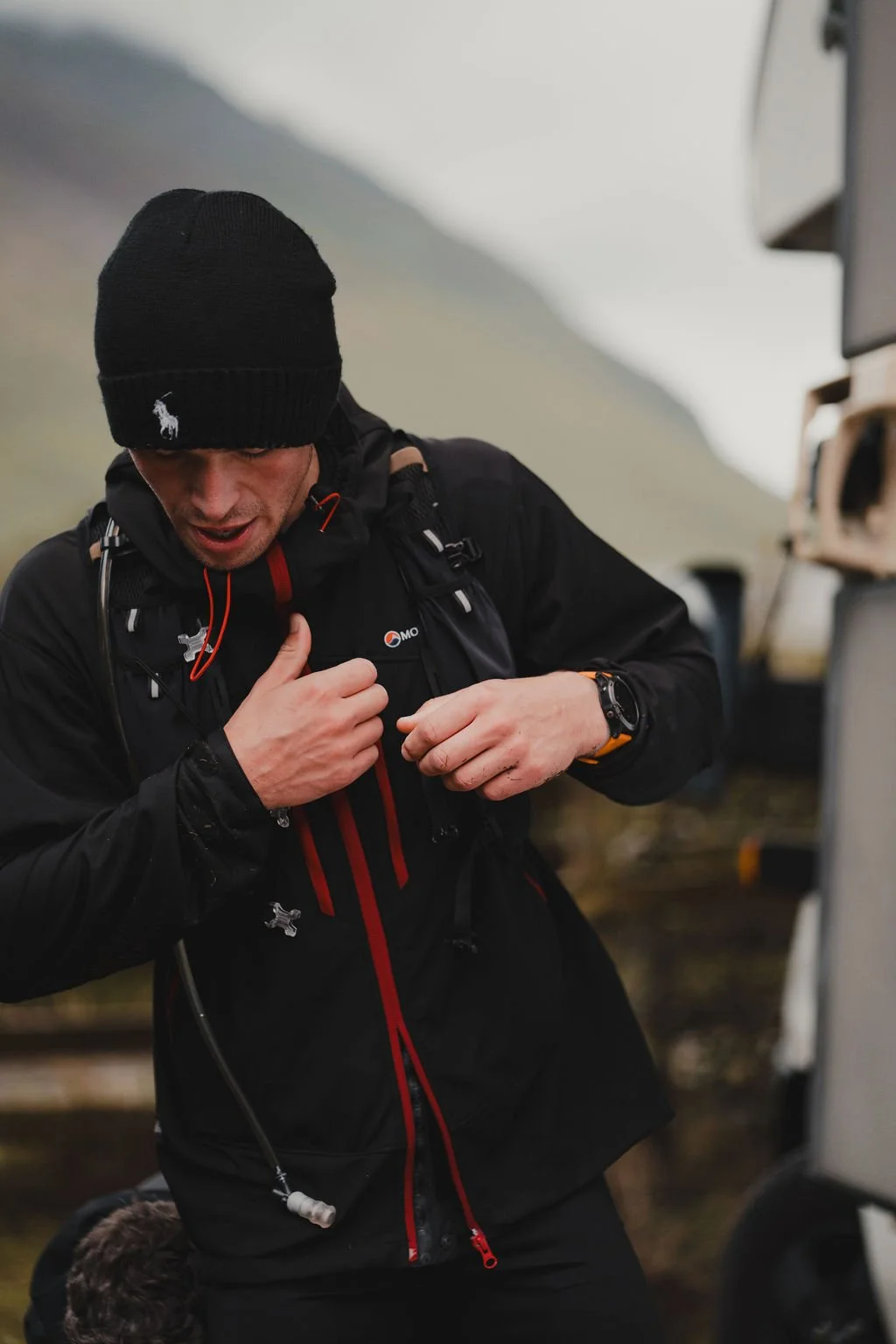 A man dressed in outdoor gear, including a black beanie and jacket, adjusting his backpack straps outdoors in a mountainous area.