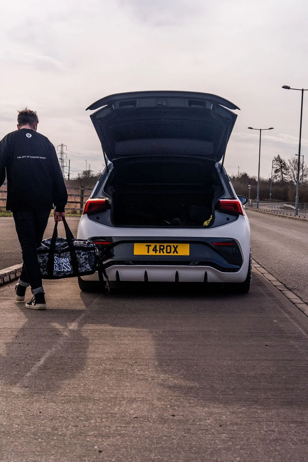 A person walking away from a white car with an open trunk, carrying a black and white duffel bag, on an empty road with a wooden fence and streetlights.