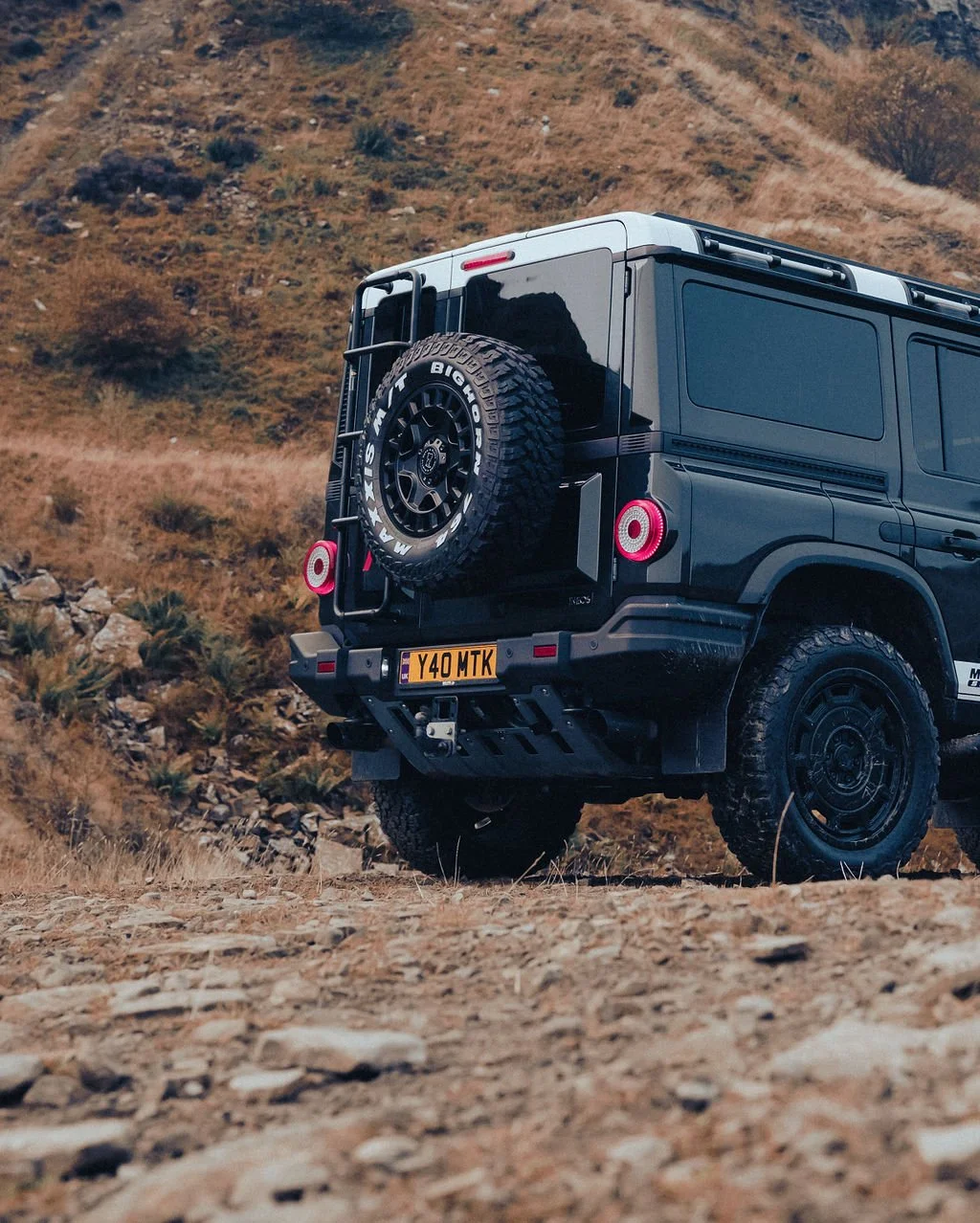 A black off-road vehicle with a rear-mounted spare tire on a rocky dirt trail in a hilly, dry landscape.