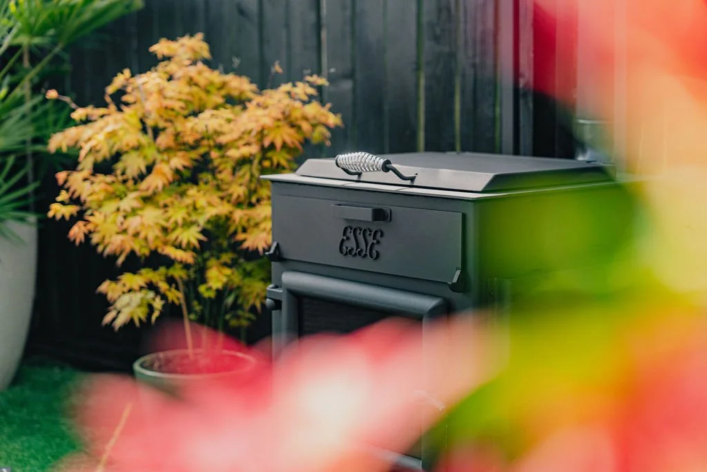 Outdoor grill on a patio with potted plants and a black fence, partially obscured by red and green foliage.