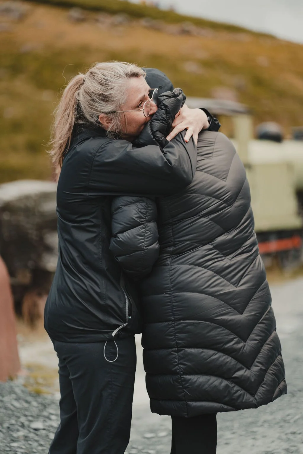 Two people hugging outdoors on a rocky surface, dressed in black jackets, with mountainous terrain and a train in the background.