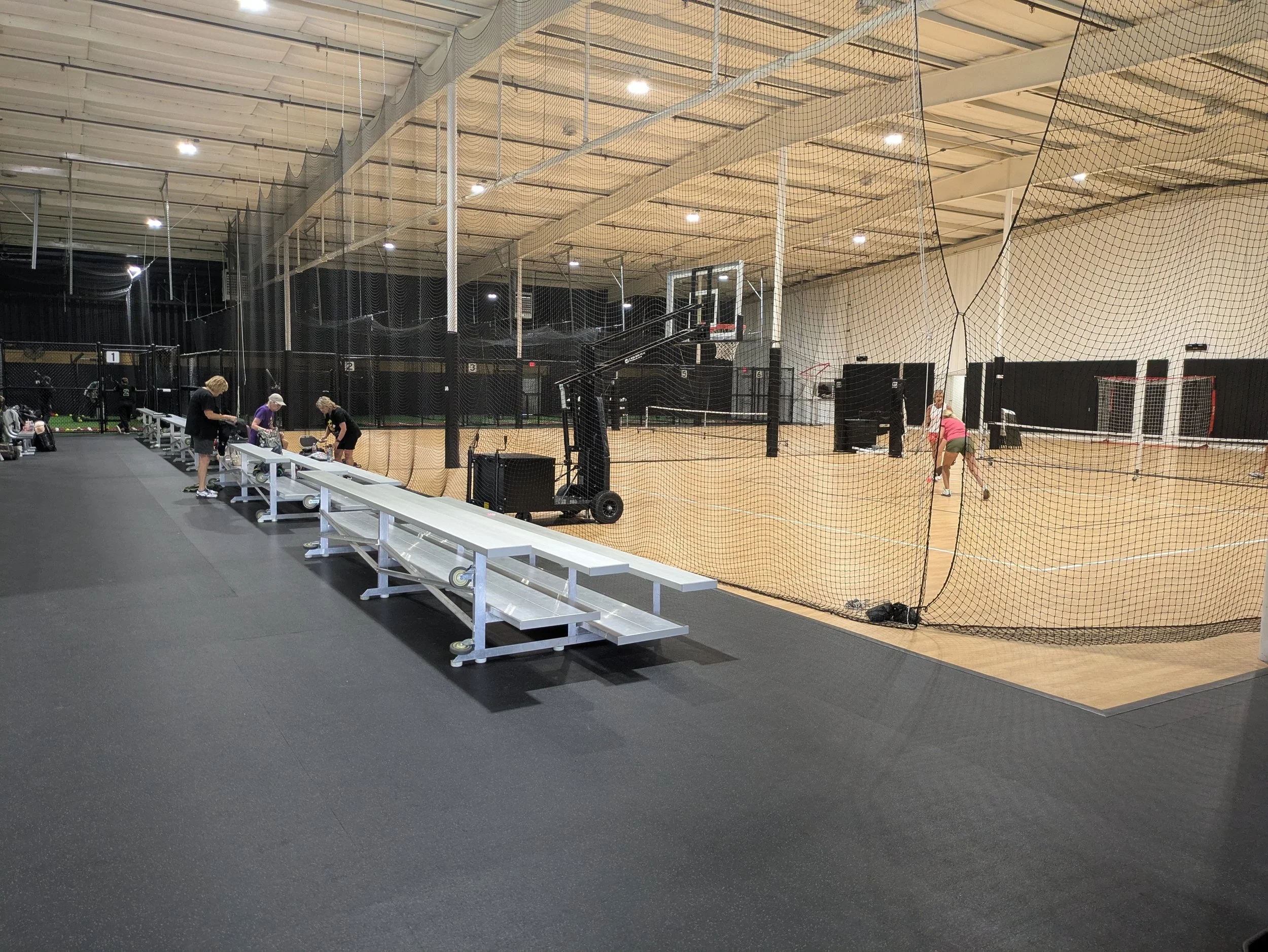Indoor sports facility with a tennis or pickleball court, net, and players practicing, separated by a safety net, with a seating area and people preparing nearby.