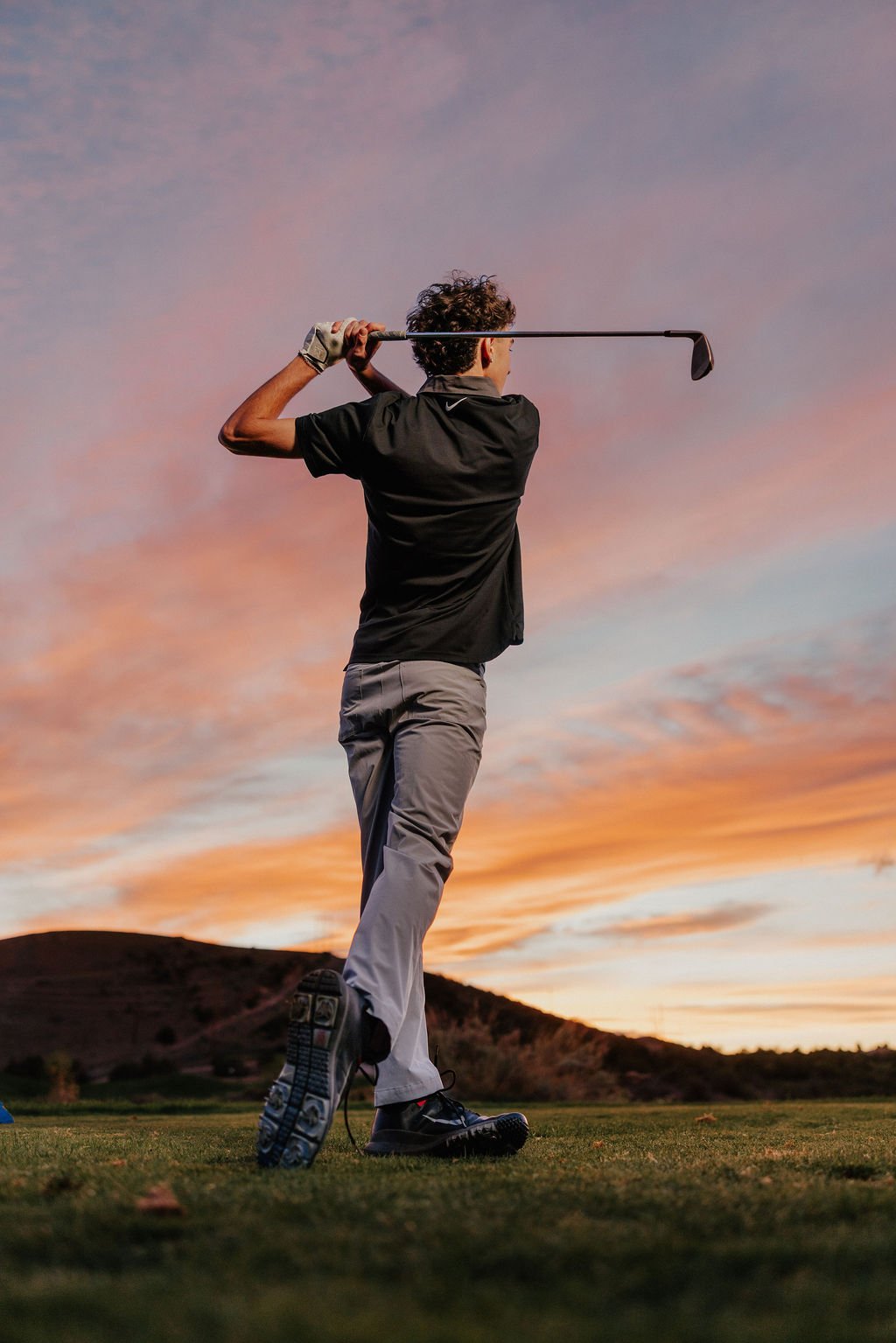 Person playing golf at sunset, swinging a golf club on a grassy field with hills in the background.