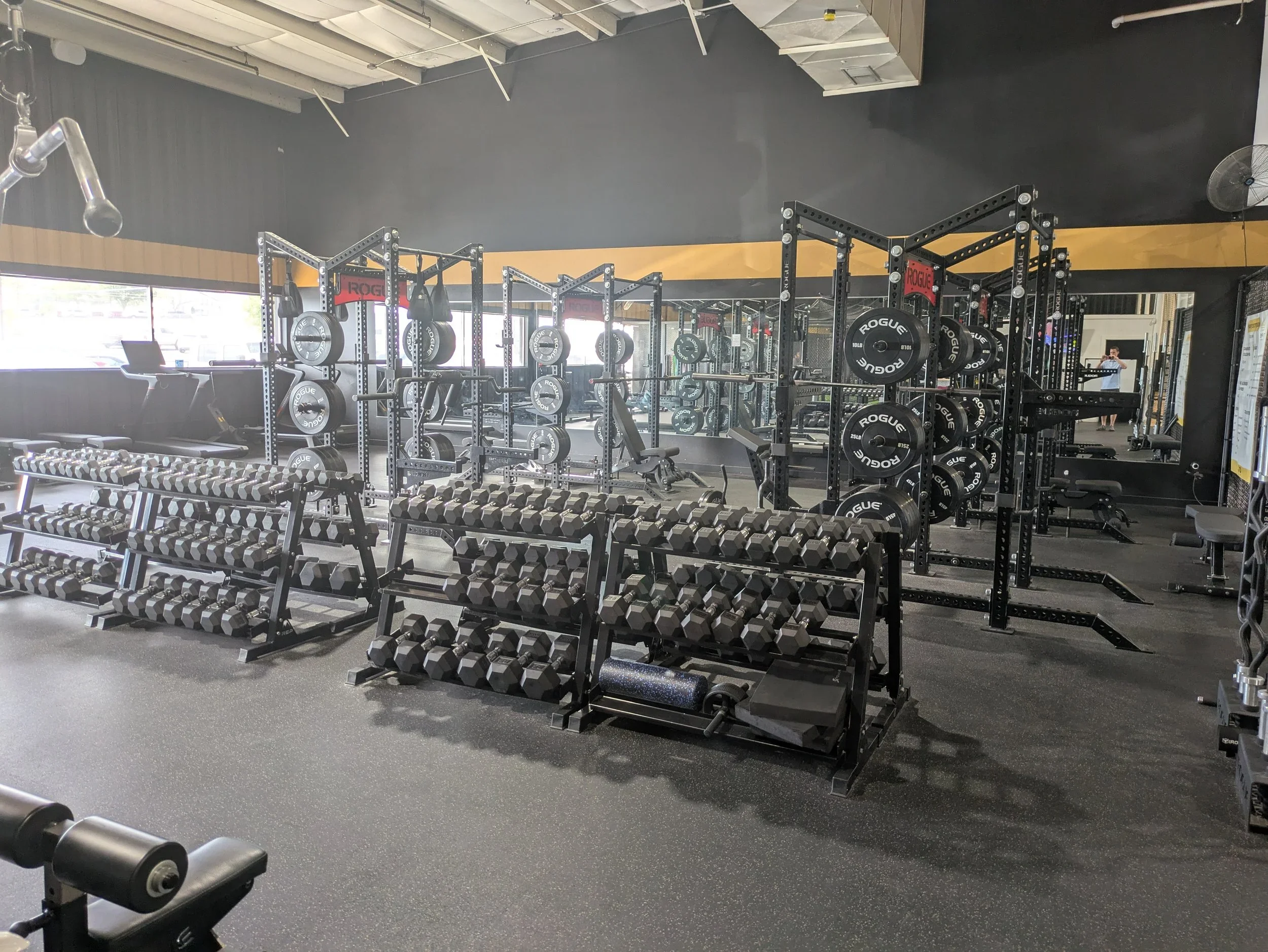 Interior of a gym with weightlifting equipment including dumbbells, barbells, and weight plates on racks, a row of mirrors, and workout benches.