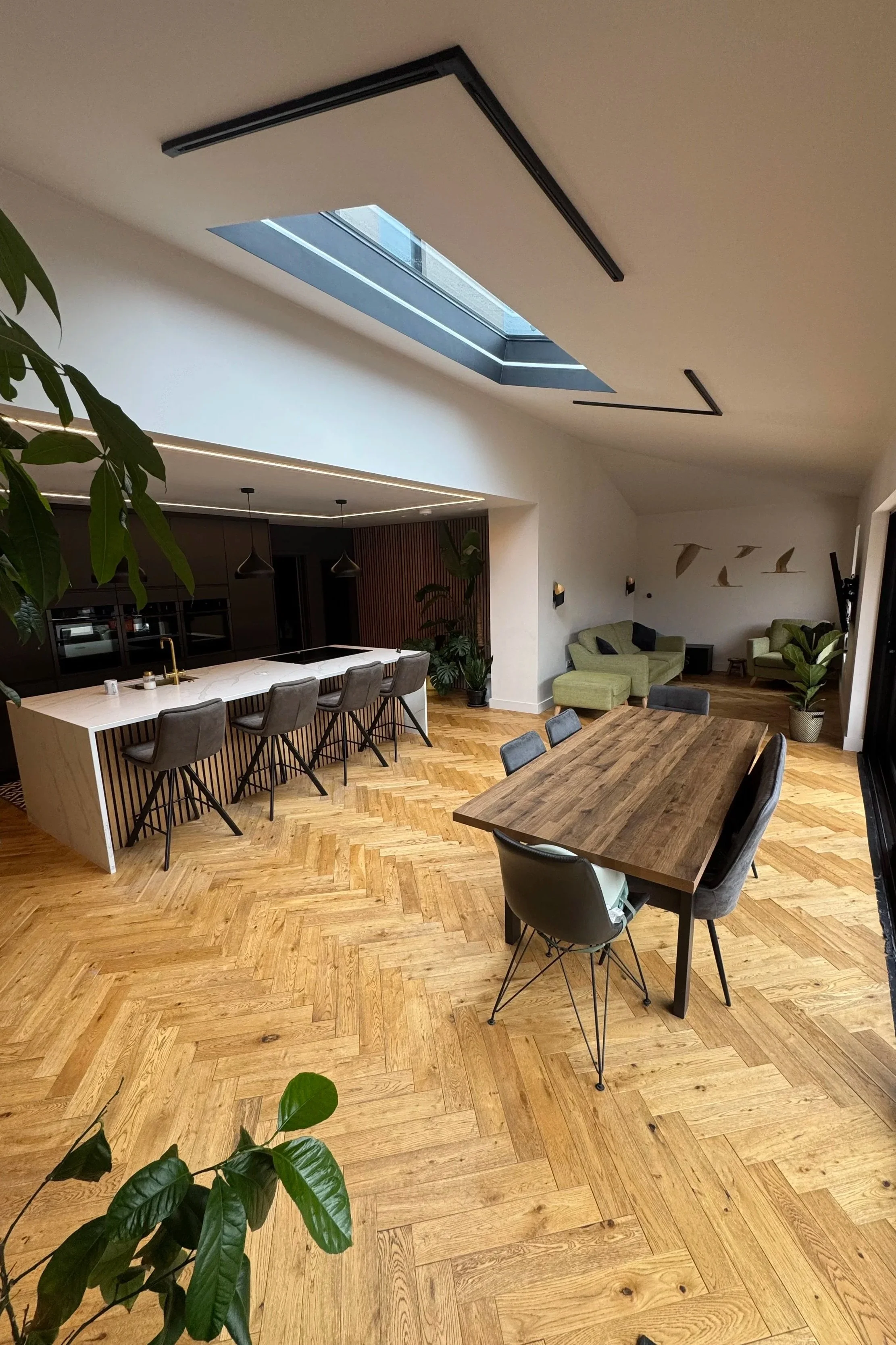 A modern, open-concept living and dining area with wooden flooring, a rectangular wooden dining table with chairs, a kitchen island with barstools, living room seating with green chairs, green plants, and a skylight in the ceiling letting in natural light.