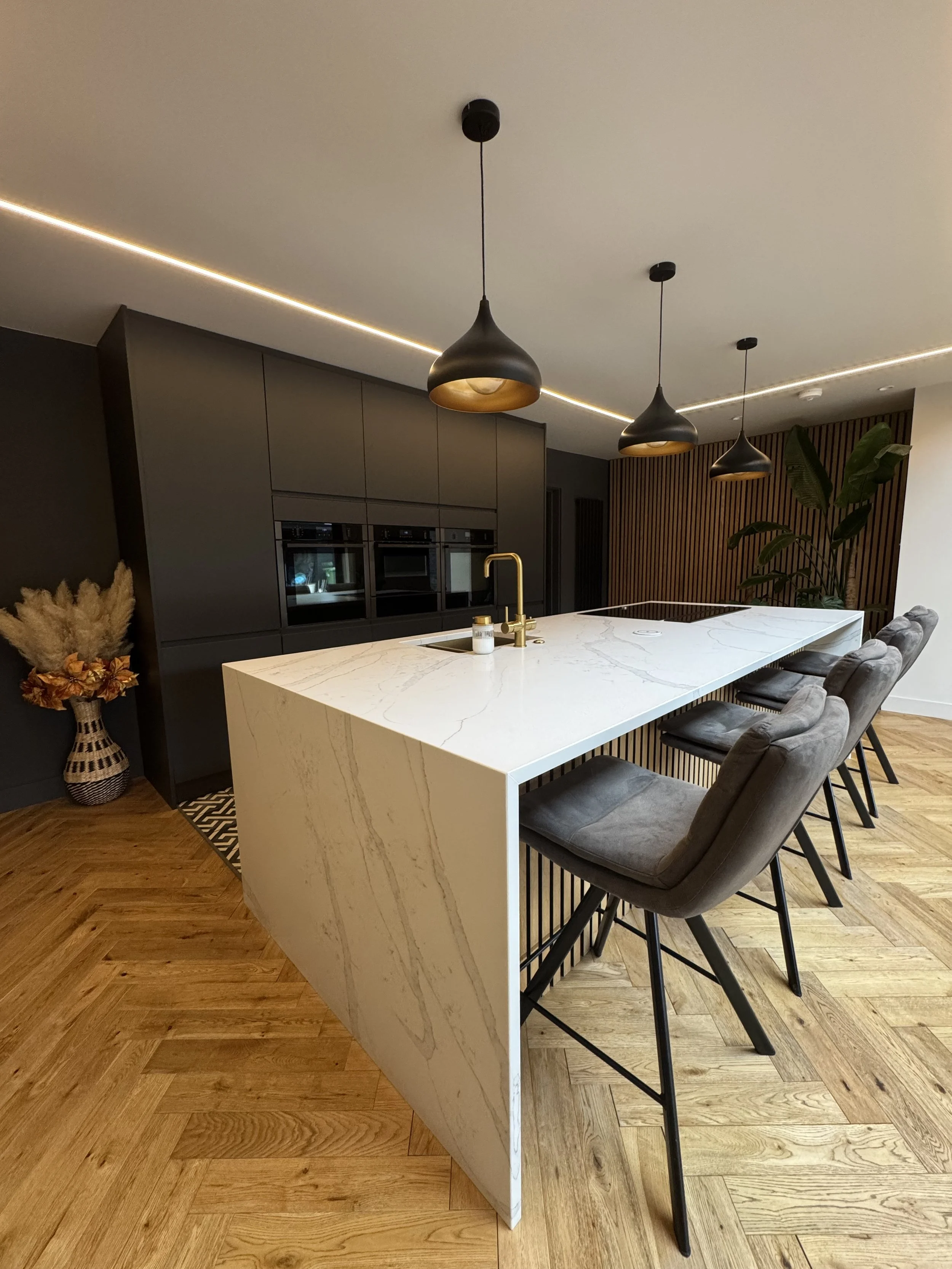 Modern kitchen with a white marble island, black pendant lights, and dark cabinetry, with a wooden herringbone floor and decorative plants.