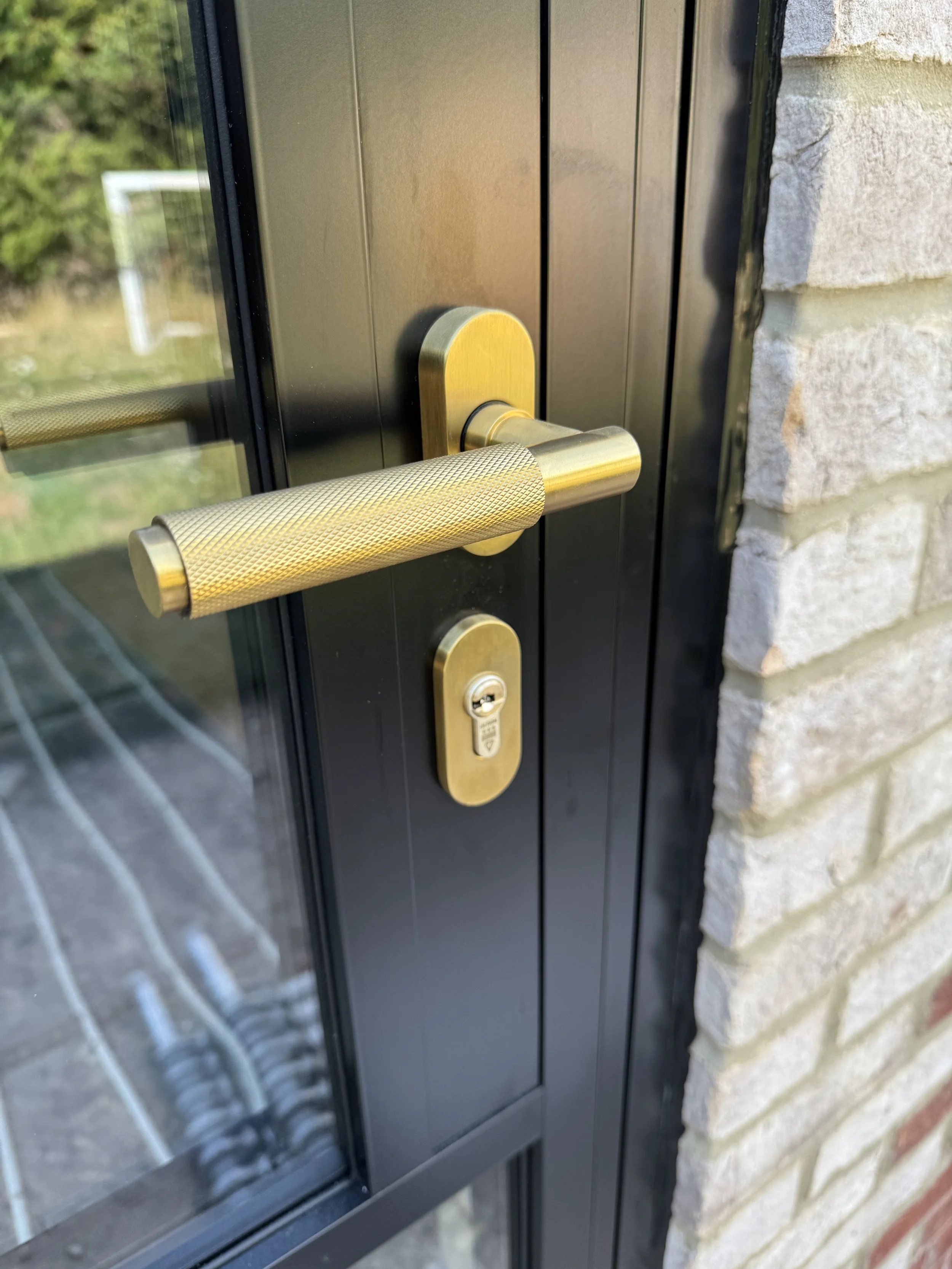 Close-up of a bespoke brass door handle and lock on a black aluminum bi-fold door against a stone wall beside it.