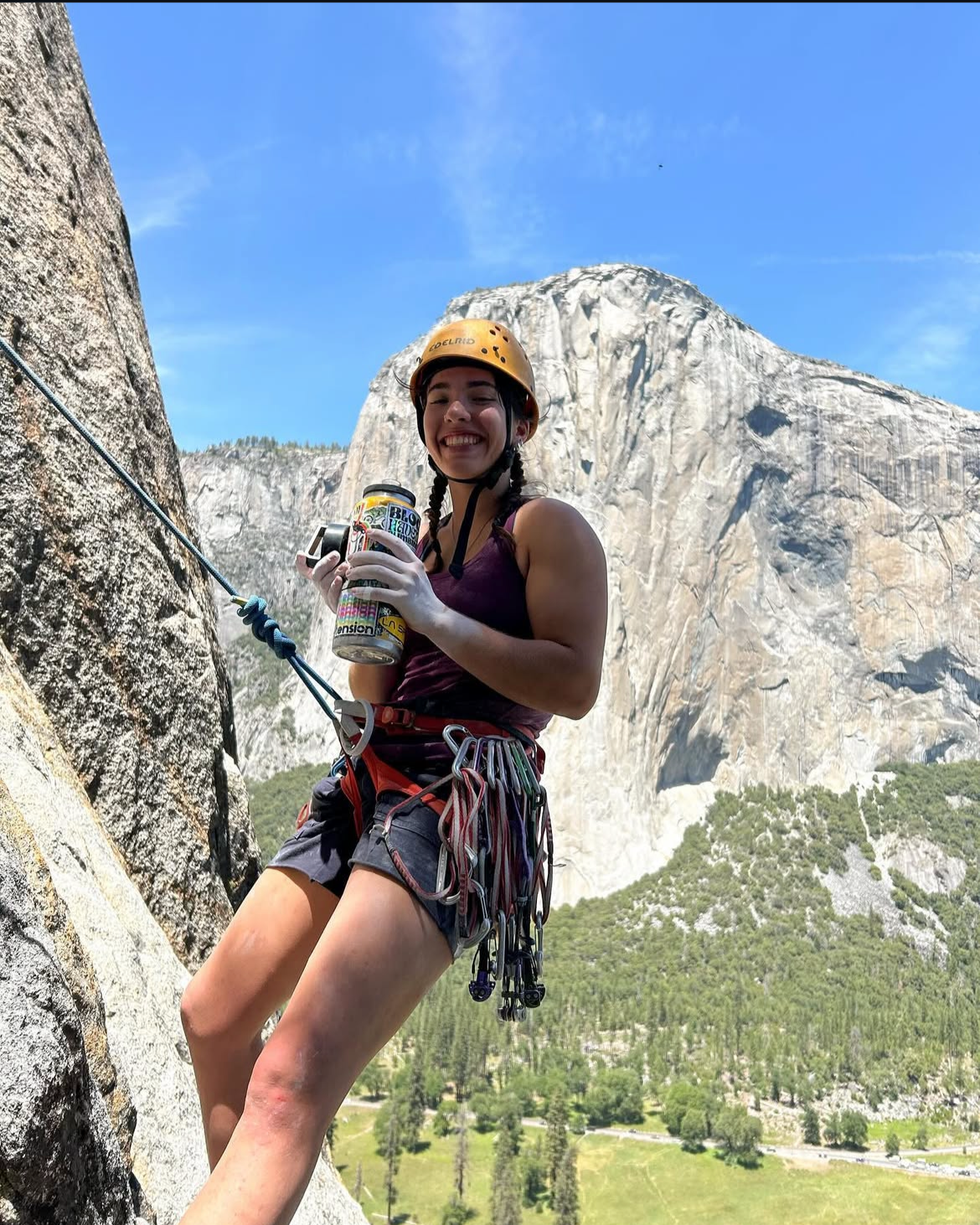 A young woman rock climbing outdoors on a steep granite wall, wearing a yellow helmet and harness, smiling and holding a drink, with a mountain and green landscape in the background.