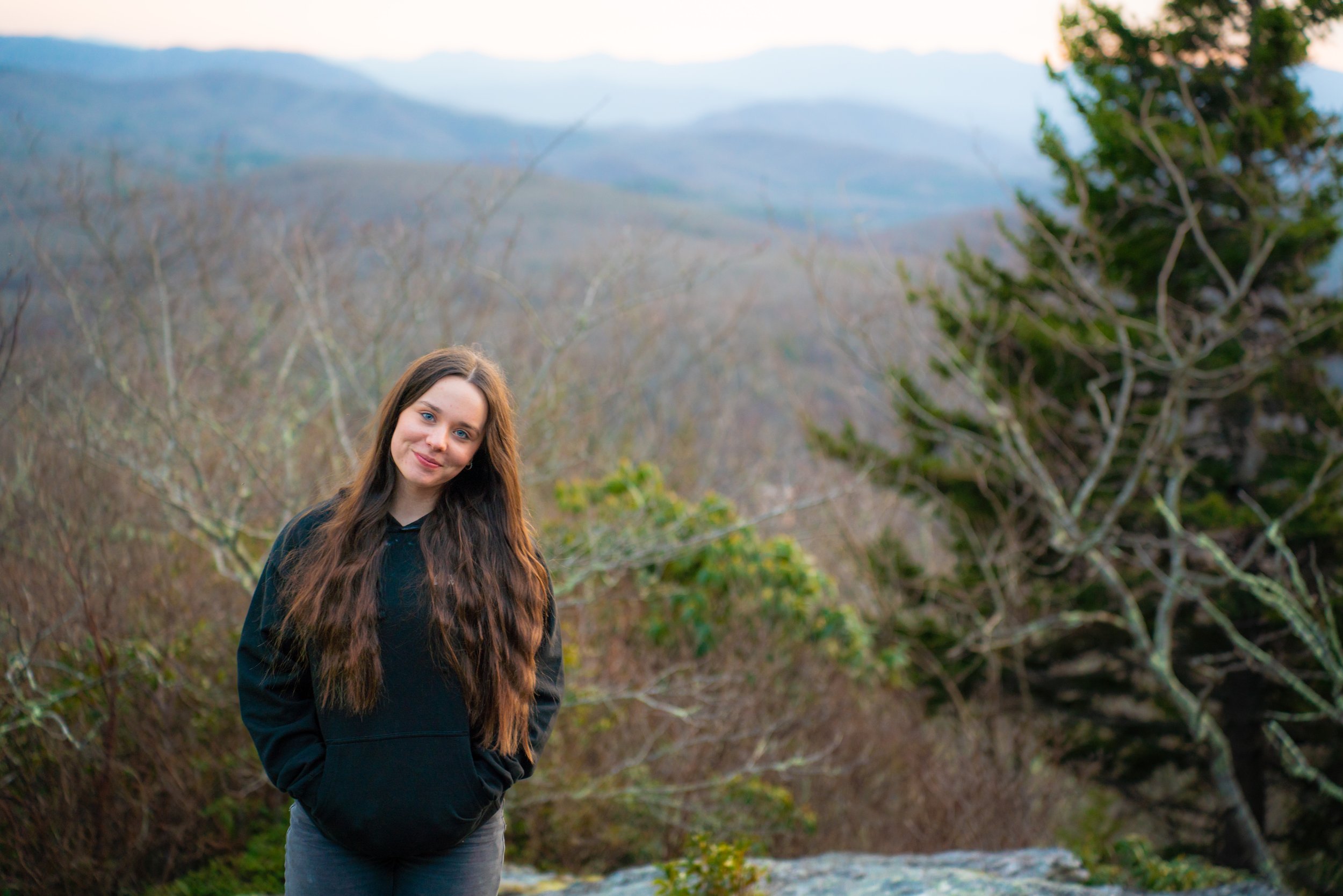 Young woman standing outdoors in front of trees and mountains, wearing a black hoodie, smiling gently with hands in pockets.