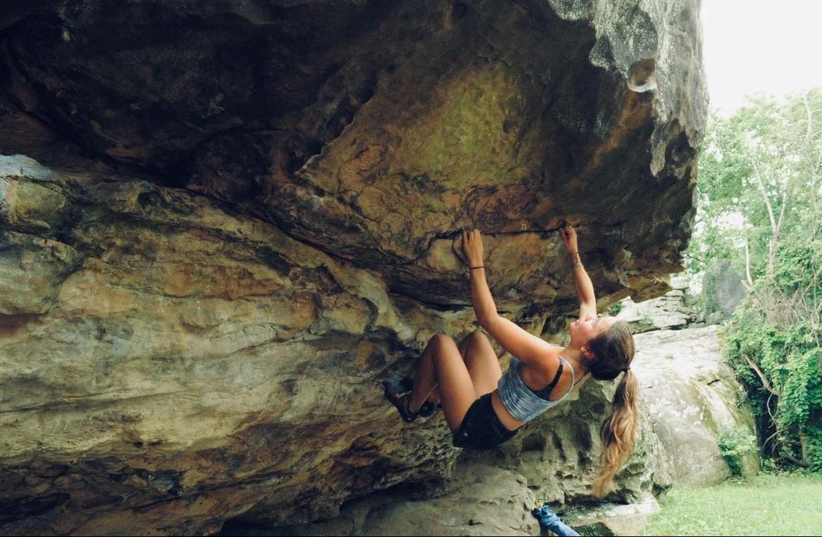 Young woman rock climbing on a natural outdoor rock formation, hanging from a horizontal crack with greenery in the background.