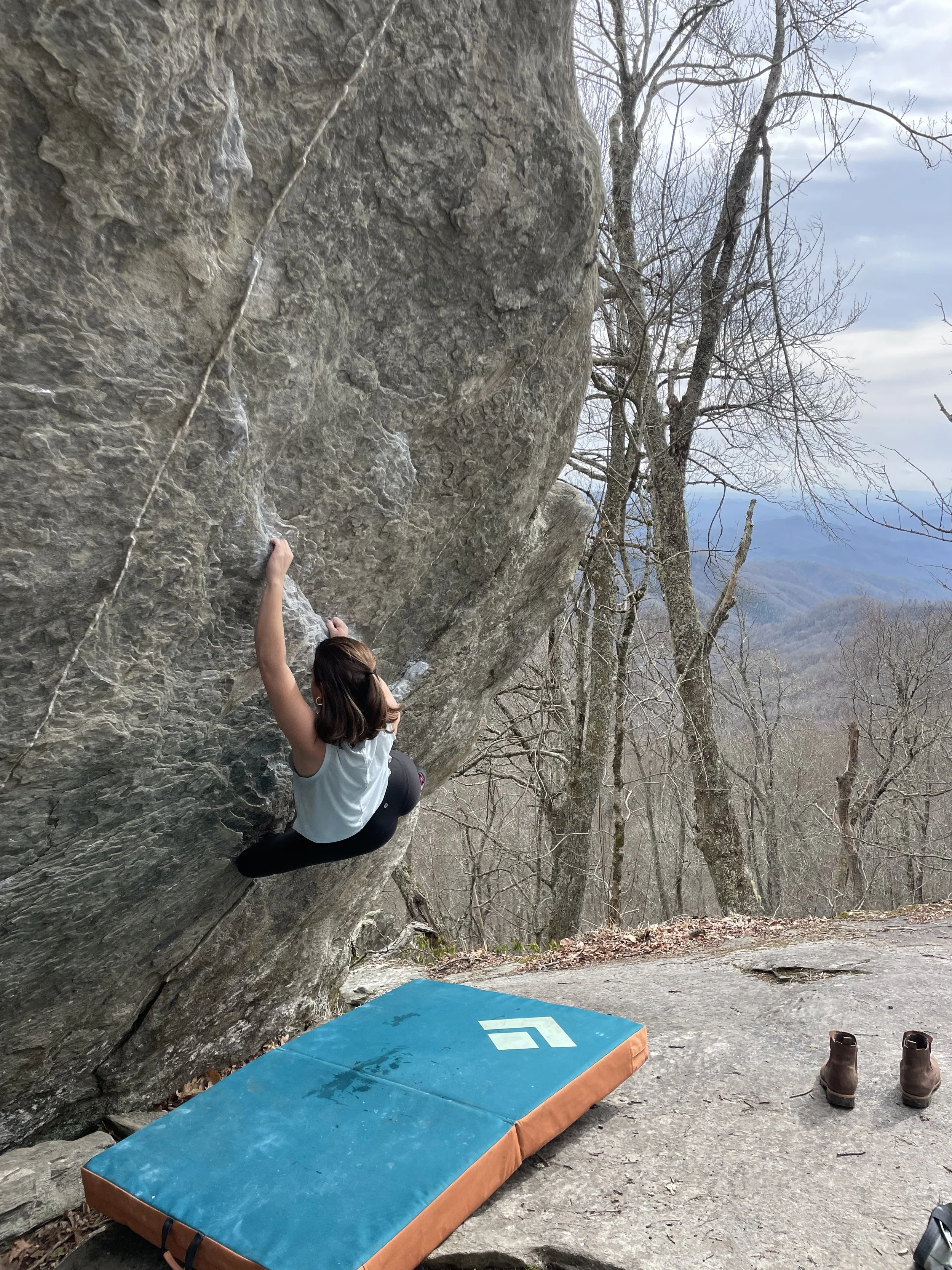A woman rock climbing on a large overhanging boulder outdoors in a wooded area, with climbing shoes on the ground nearby and a crash pad underneath the climb.