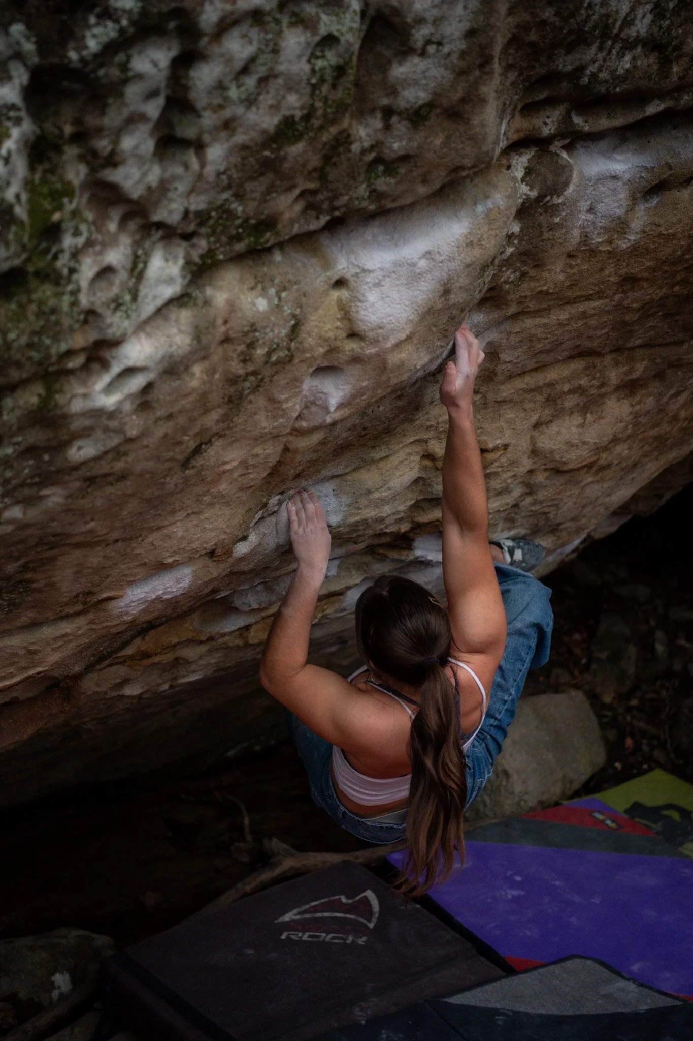 A woman with long brown hair in a ponytail is rock climbing on an overhanging rock face, with climbing mats placed below her for safety.