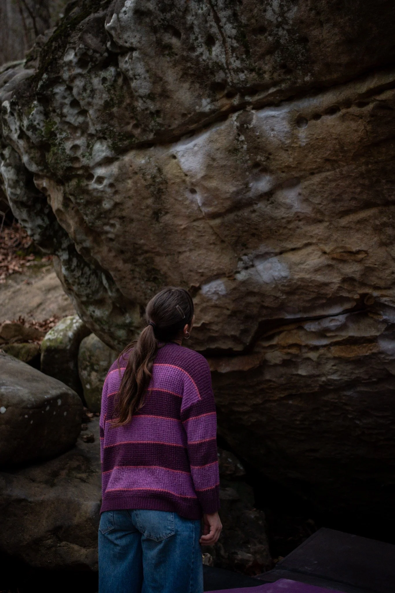 A woman with long brown hair tied back, wearing a purple and pink striped sweater and blue jeans, standing outdoors in front of large rocks in a natural setting.