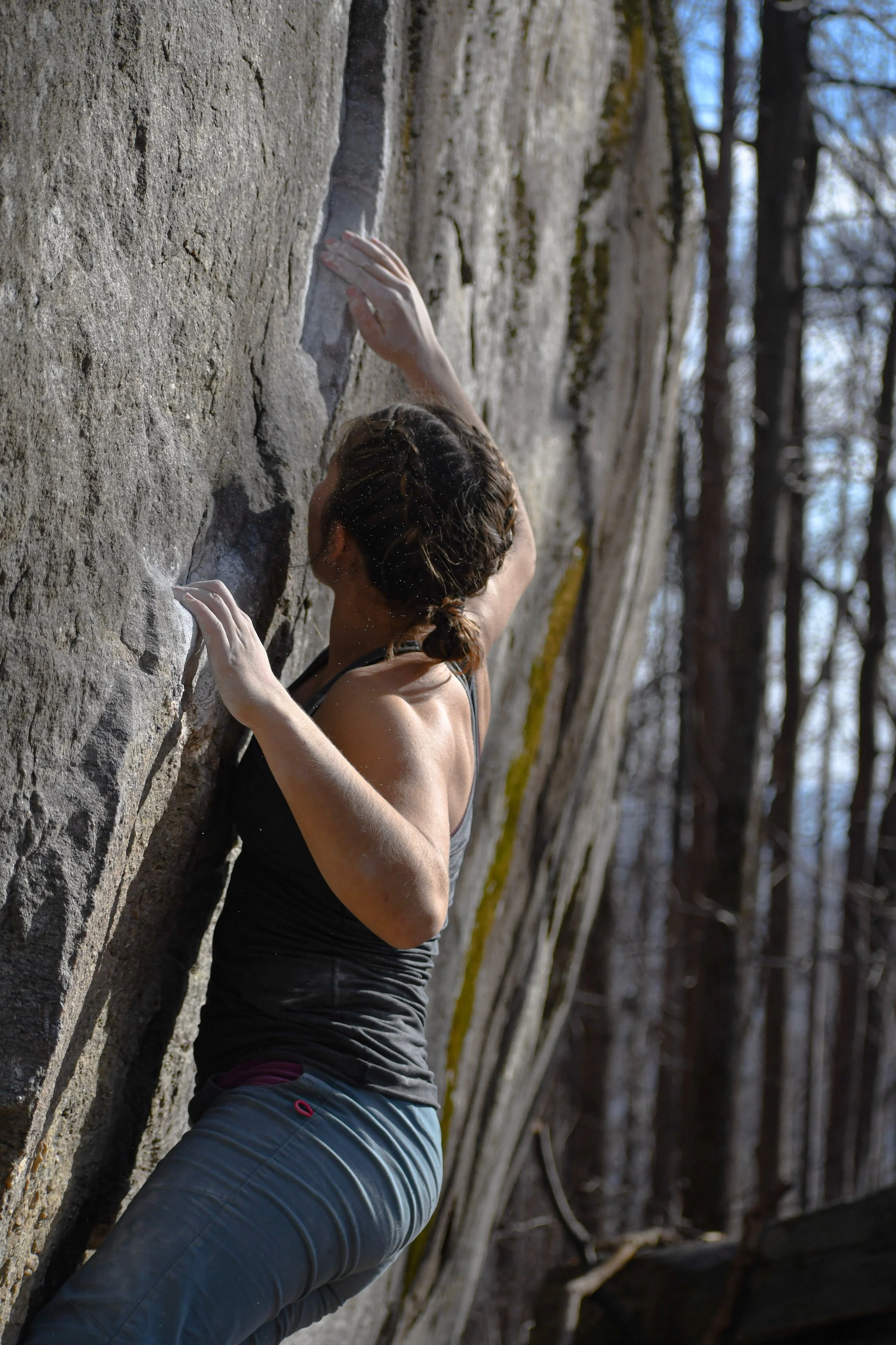 A woman rock climbing outdoors on a vertical rock wall, wearing a black tank top and gray pants in a wooded area.