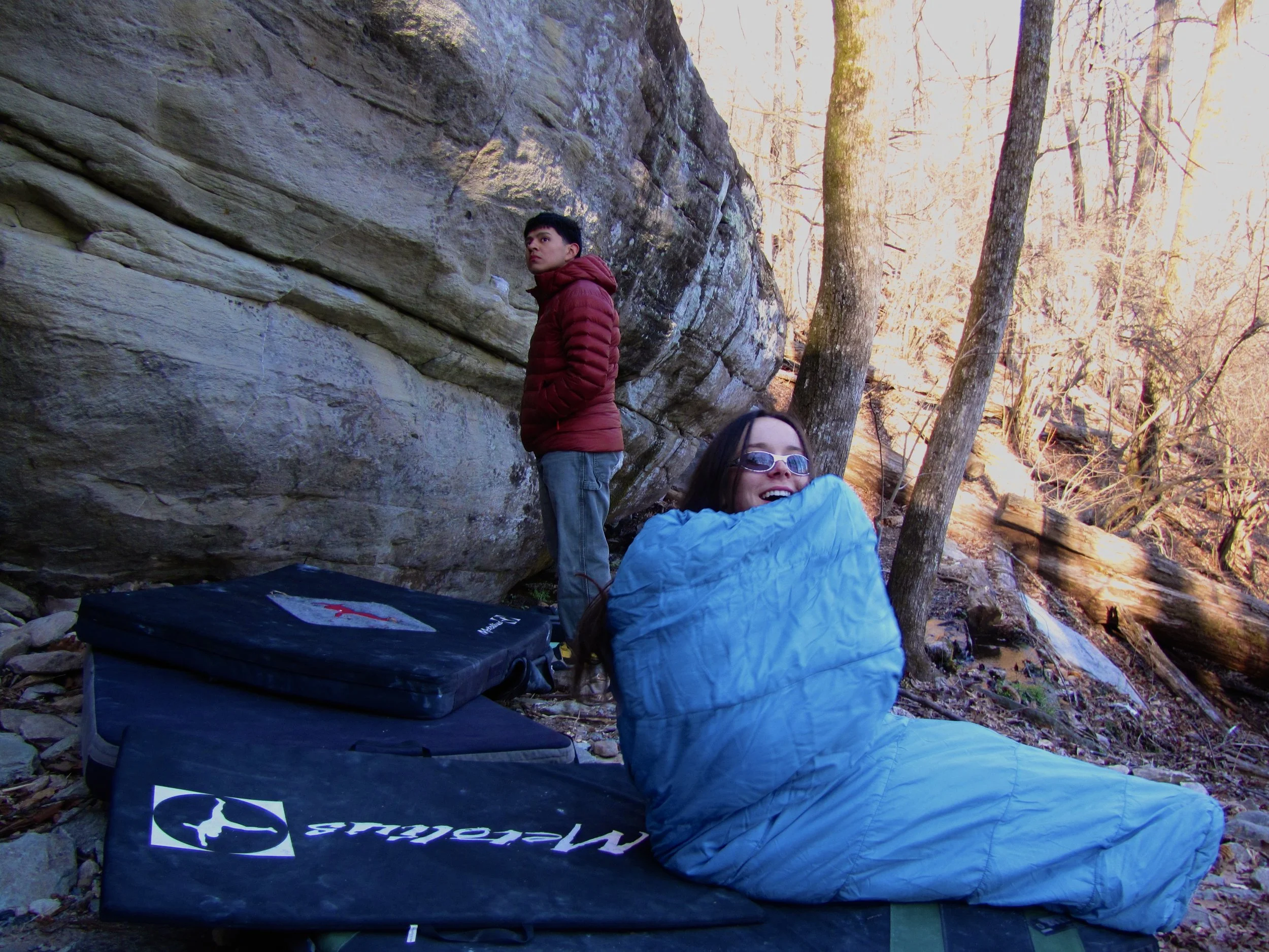 A woman sitting on a blue sleeping bag outdoors in a wooded area, smiling, with a man standing nearby leaning against a large rock, during daytime.