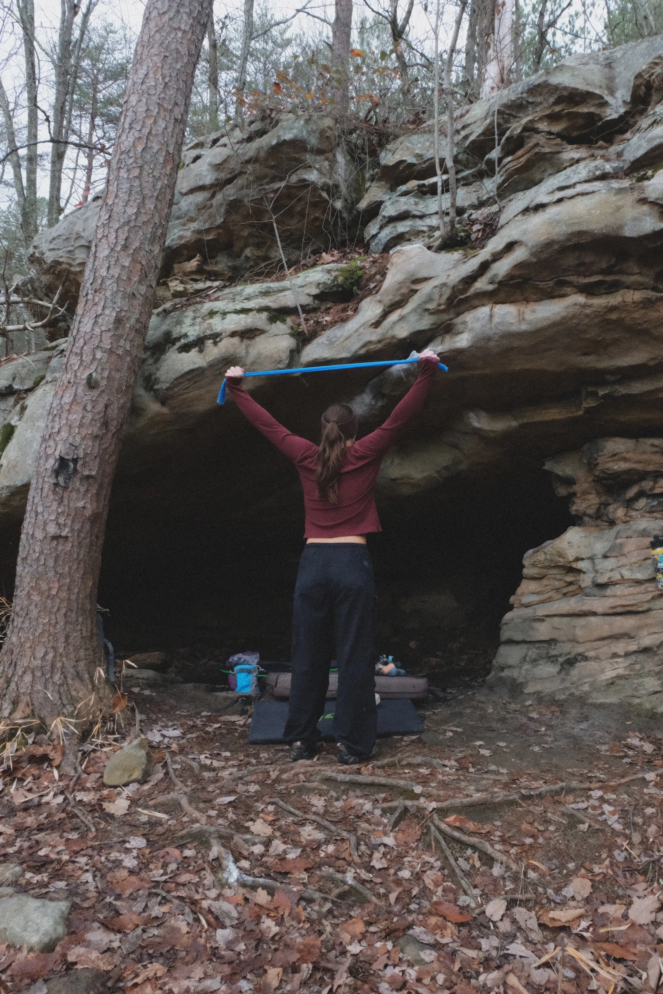 Person stretching with a stick in front of a rocky cave entrance in a wooded forest.