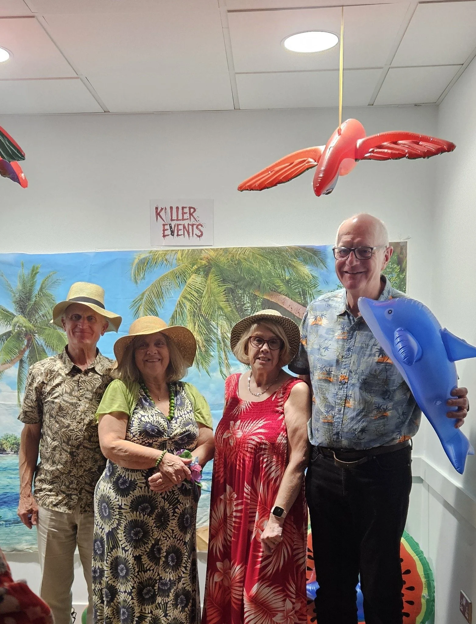 Group of four seniors in tropical-themed clothing and hats, standing in front of a beach scene backdrop, with colorful inflatable fish and a sign that reads "Killer Events," in a room decorated with tropical and beach-themed decor.