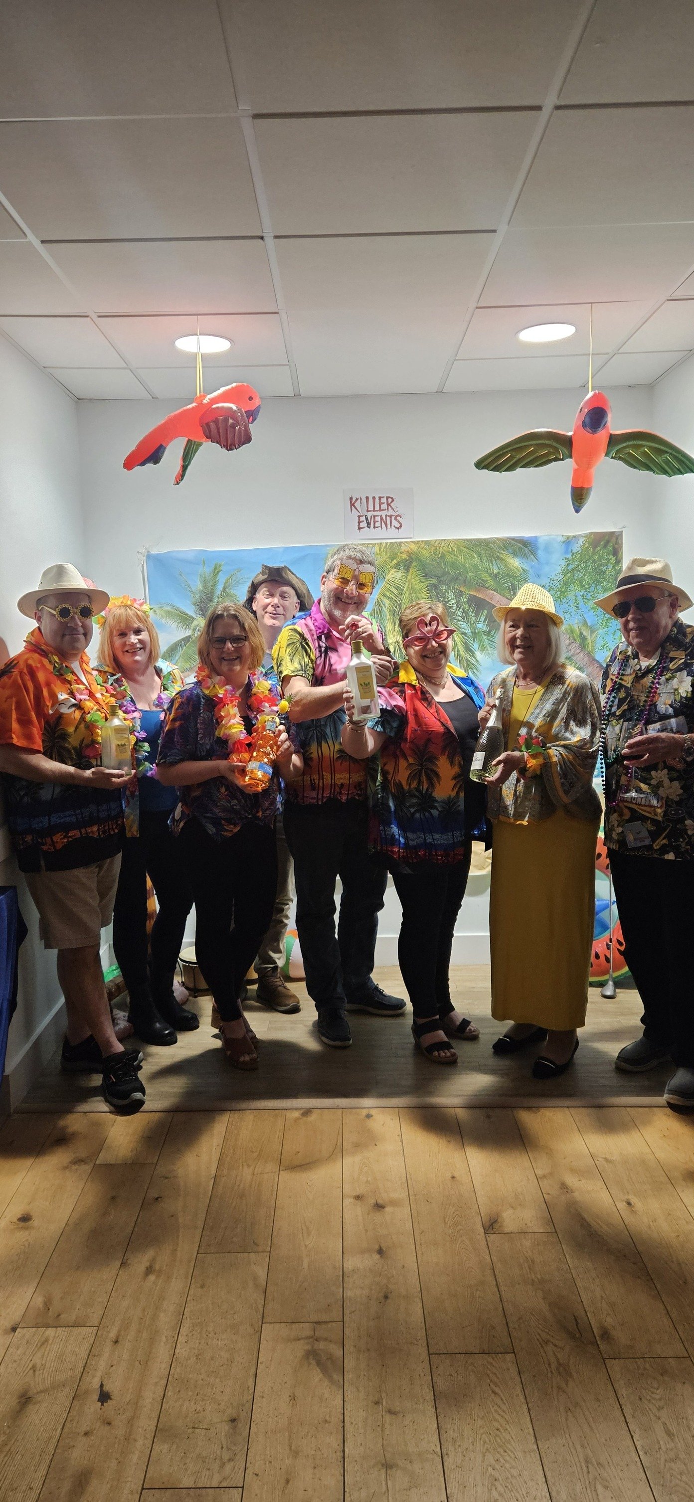 Group of people dressed in colorful tropical-themed shirts and party accessories, standing in front of a tropical backdrop with palm trees, celebrating a party with drinks and leis.