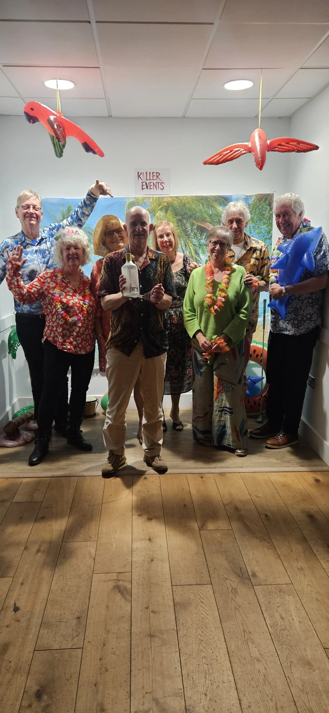 Group of people at a tropical-themed party with balloons and decorations, including flamingos and a piñata, in a room with a wooden floor and a blue tropical backdrop.