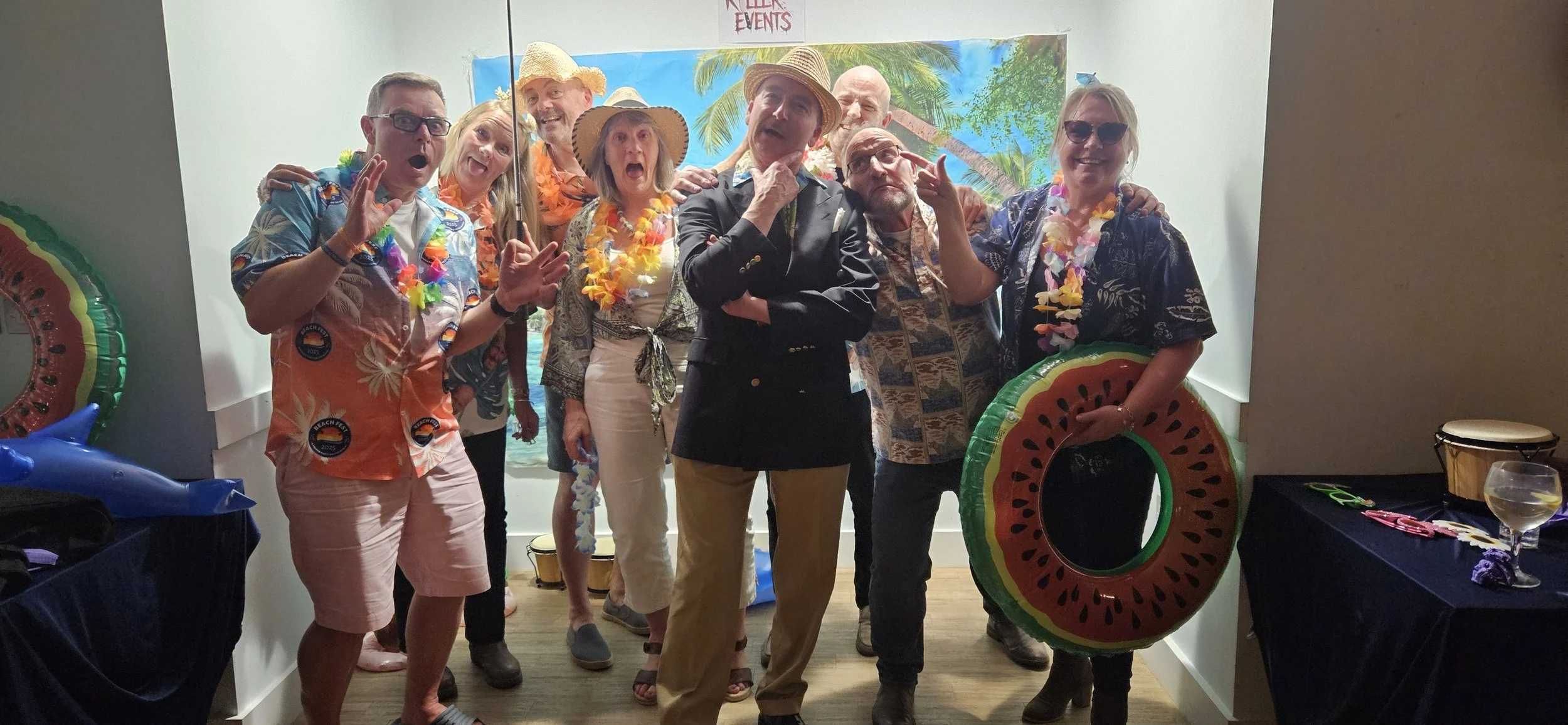 Group of people dressed in tropical shirts and leis, some wearing straw hats, posing in front of a tropical-themed backdrop at a summer party or celebration.