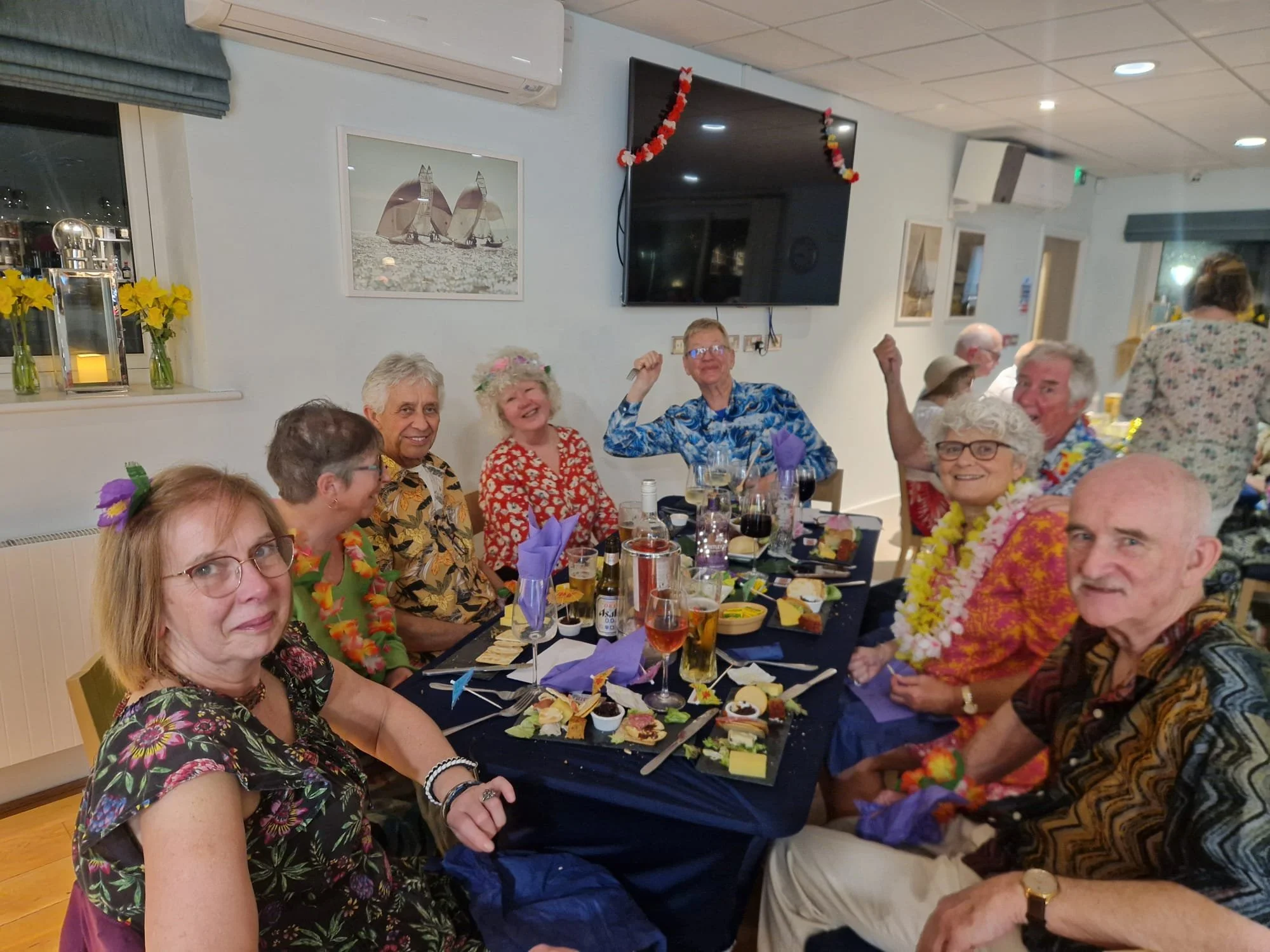 A beige speckled ceramic vase with a narrow neck and wider base, holding dried branches with small round seed pods, placed on a wooden surface with shadow cast on a light-colored wall.