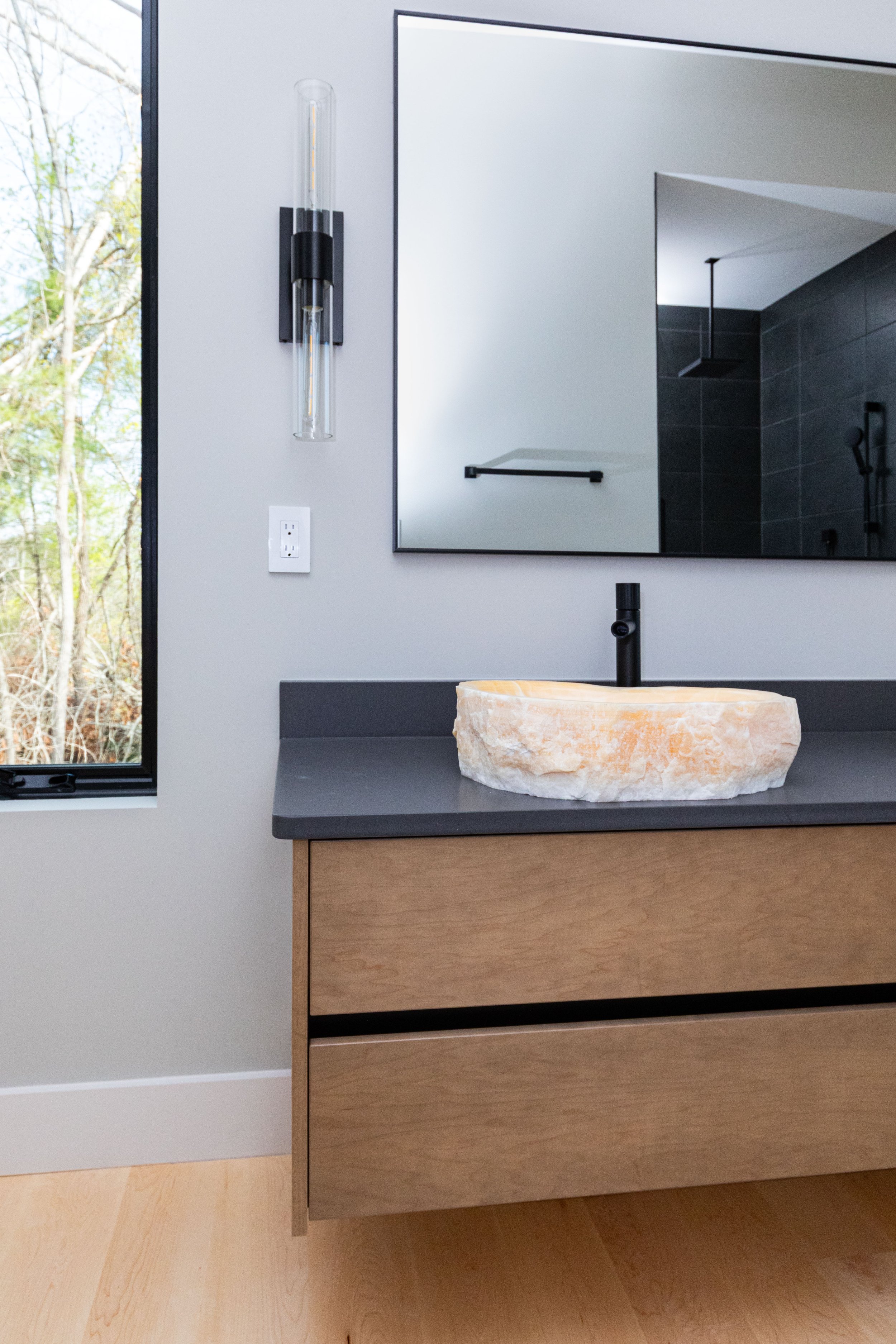 Modern bathroom with a stone vessel sink on a black countertop, a large mirror, a black faucet, a sleek black wall-mounted light fixture, and a view of a wooded outdoor scene through a window.