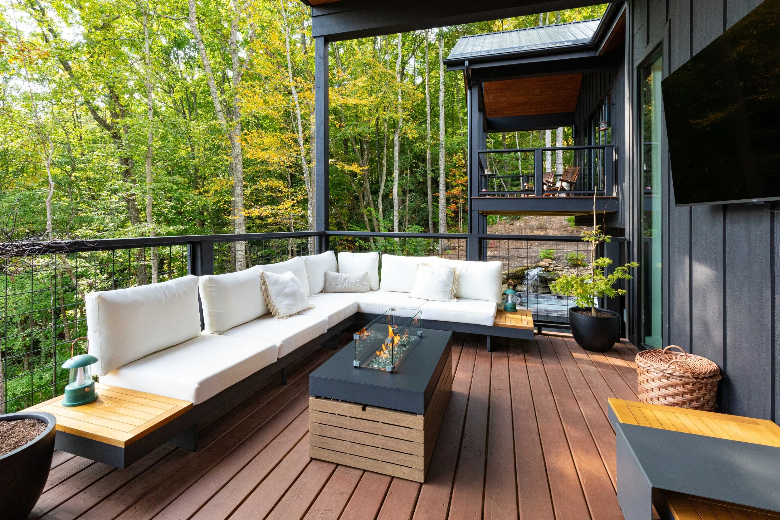 Outdoor balcony with white cushioned sectional sofa, wooden tables, potted plants, and a fire pit, overlooking a wooded backyard with trees and additional balcony above.