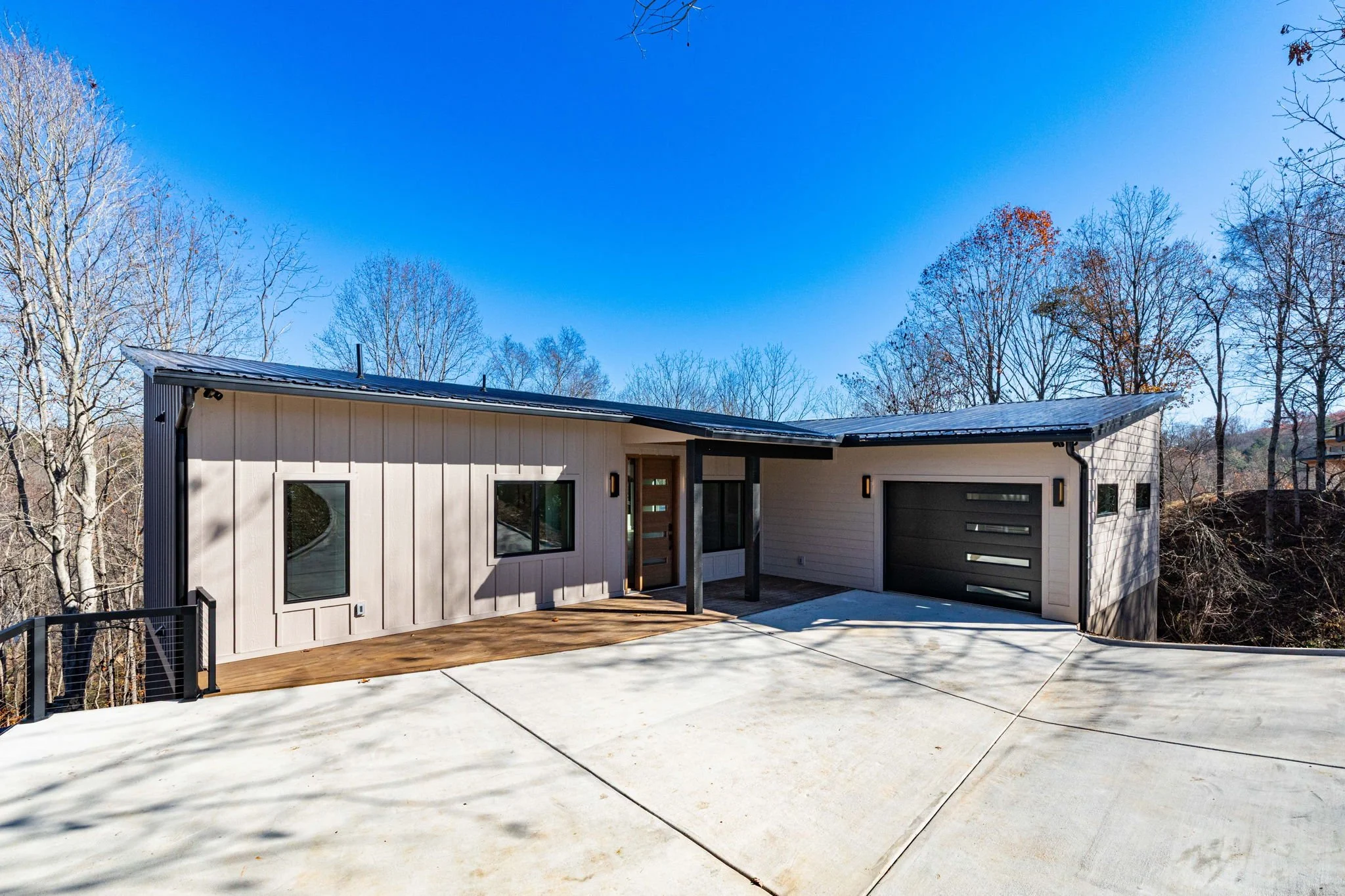 Modern house with a flat wooden porch, large windows, and a black garage door, surrounded by leafless trees and a clear blue sky.