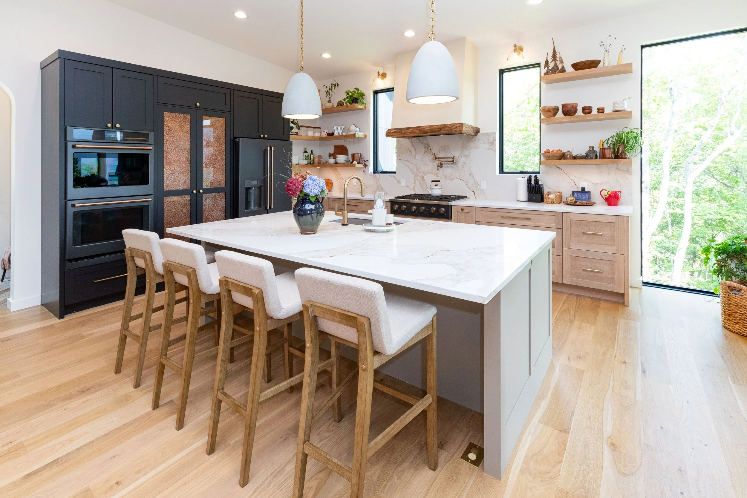 Modern kitchen with black cabinets, white marble island, light wood lower cabinets, and open shelves, with large windows and hardwood floors, decorated with plants and pottery.