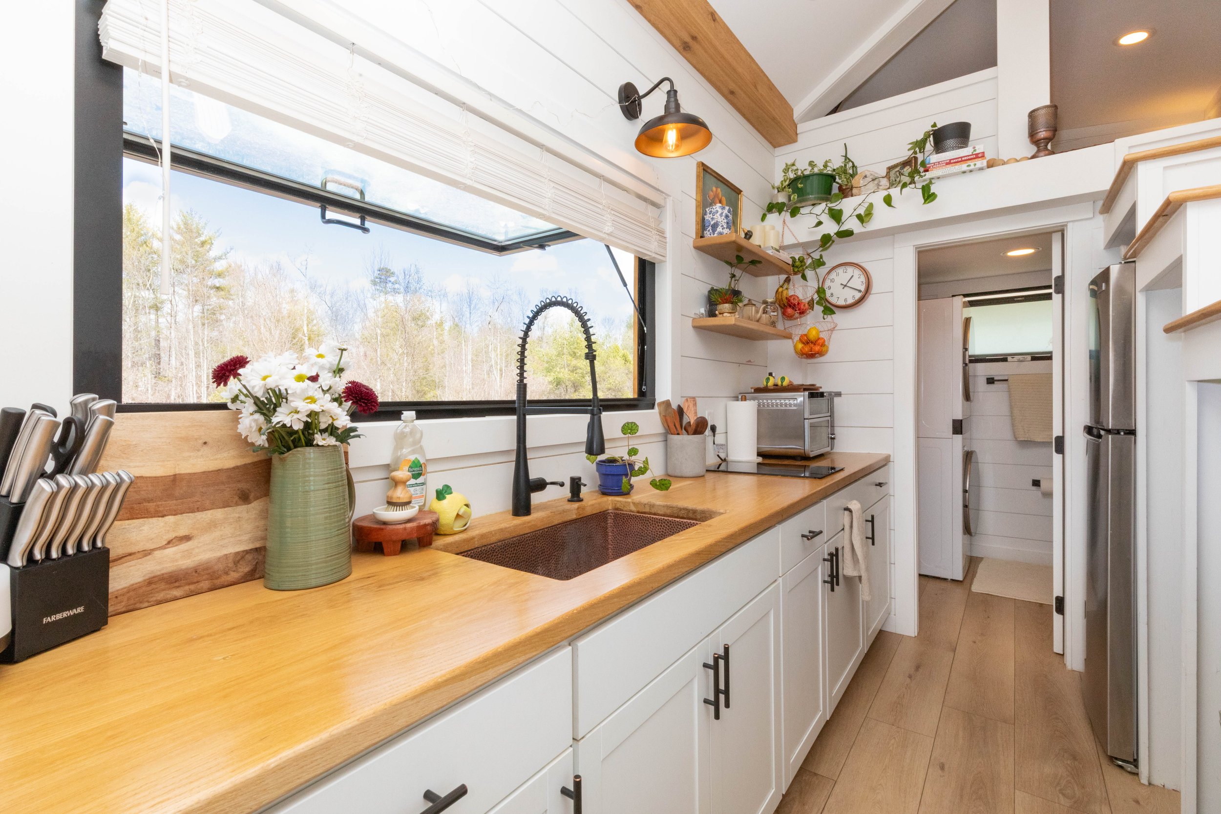A compact kitchen with a large window above the sink, wooden countertops, white cabinets, and open shelves decorated with plants and kitchen items. There's a vase of flowers on the counter and a small mural in the back of the kitchen with a doorway leading to a laundry area.