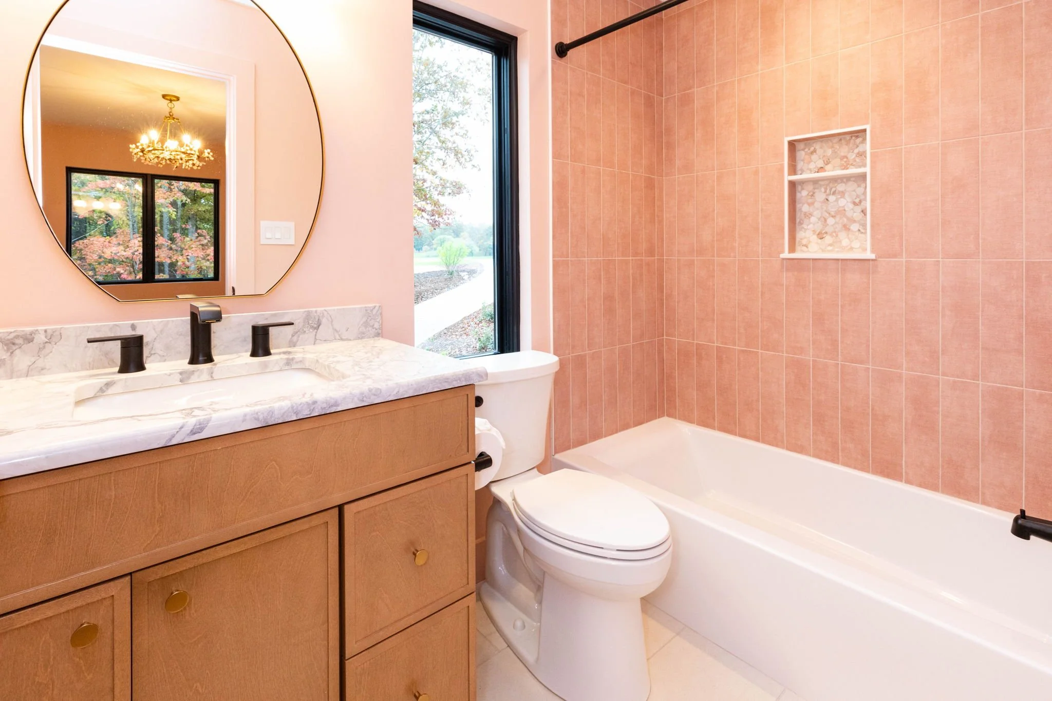 Bathroom with peach-colored tiles around bathtub, a window showing an outdoor view, a white toilet, wooden vanity with marble countertop, oval mirror, and a chandelier in the adjacent room reflected in the mirror.