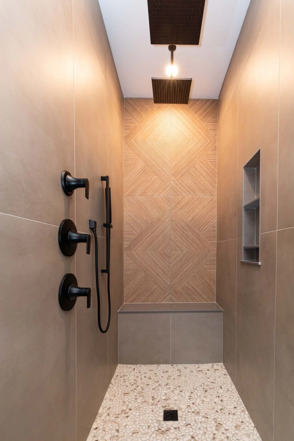 Modern walk-in shower with black fixtures, handheld showerhead, overhead rain shower, beige tiled walls, built-in shelf, and small pebble floor.
