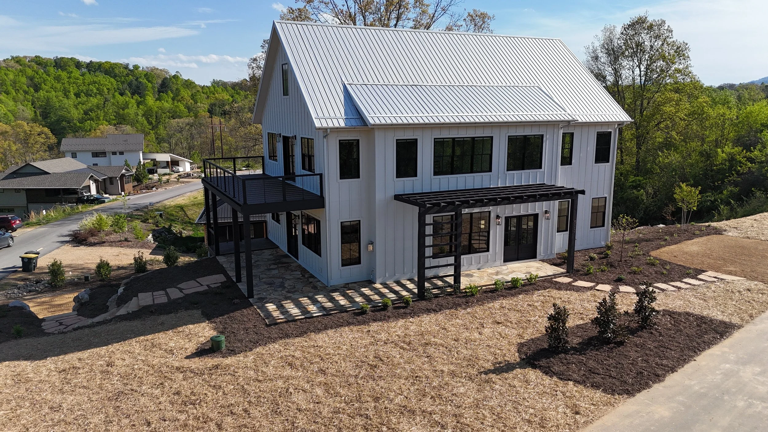 A modern two-story house with a white exterior, black window frames, and a metal roof, featuring balconies and a landscaped yard with small plants and an uneven stone pathway.
