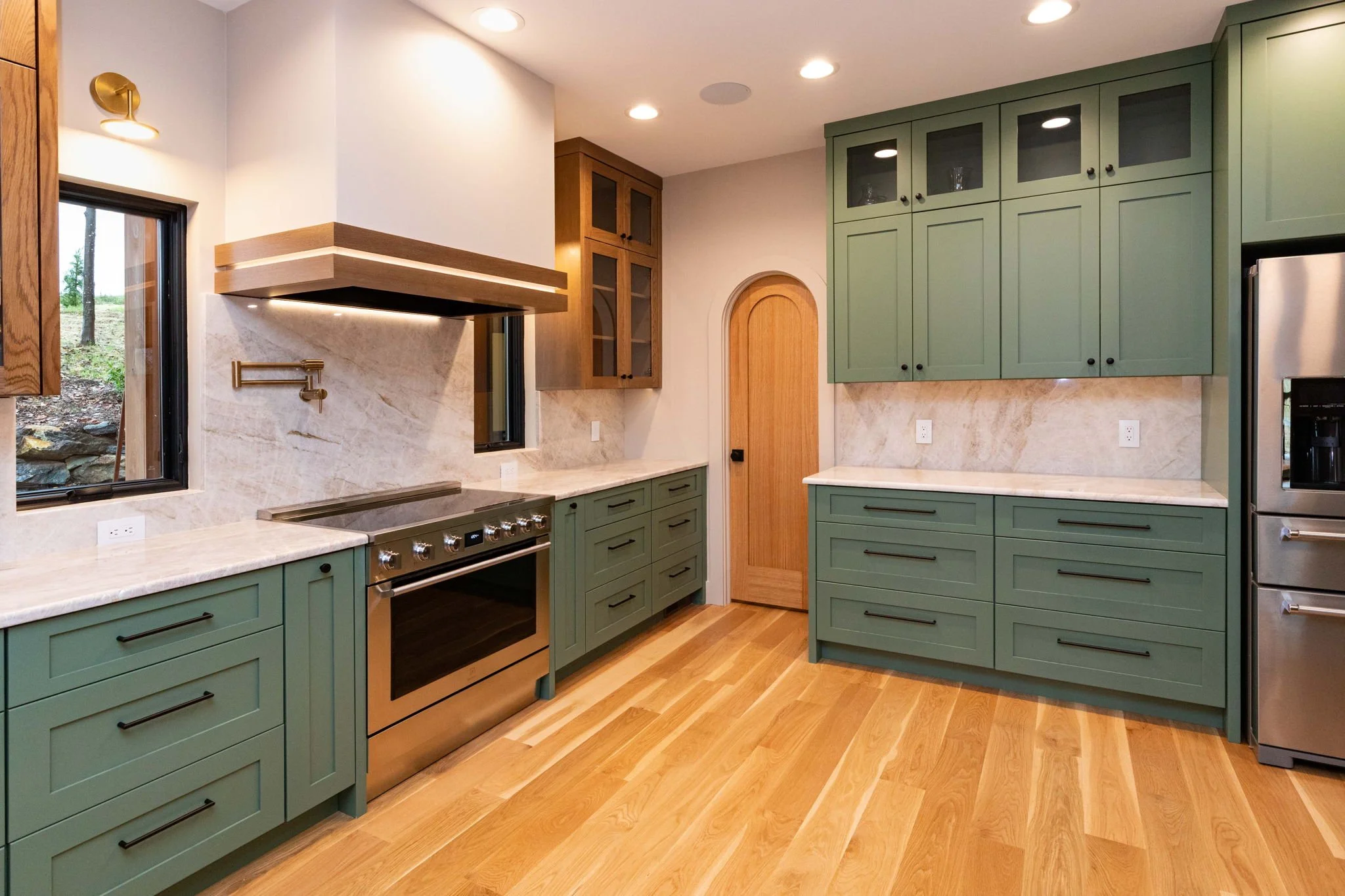 Modern kitchen with green cabinets, a stainless steel oven, marble countertops, light wood flooring, and natural light from windows.