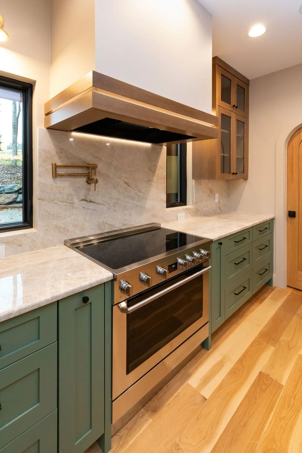 Kitchen with green cabinets, a stainless steel stove, marble countertops, a wooden range hood, and upper wooden cabinets with glass doors.