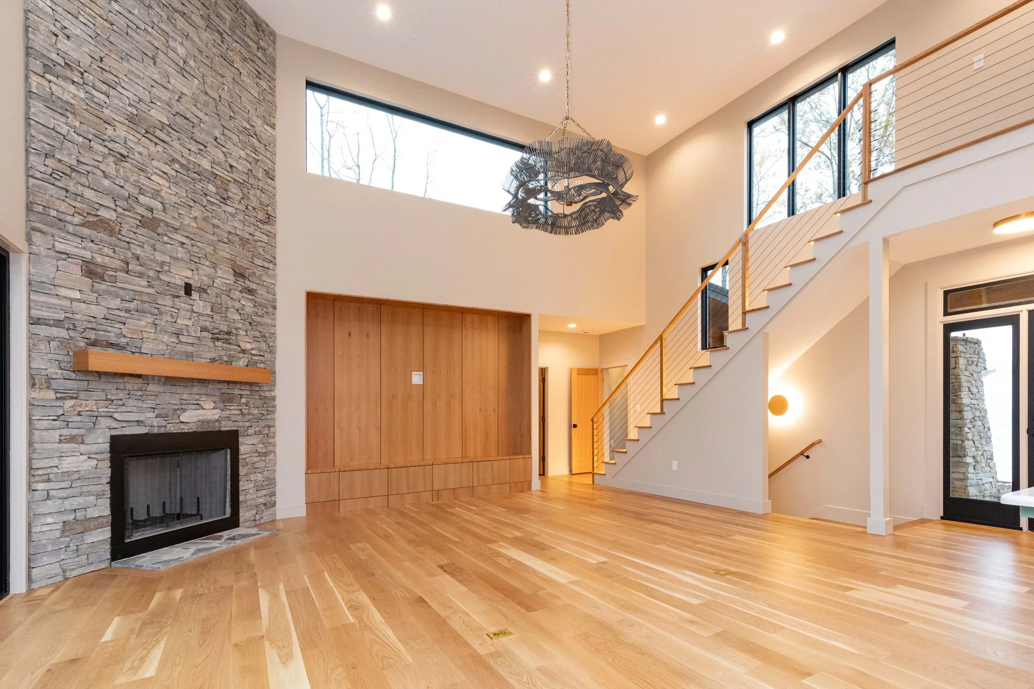 Bright modern living room with high ceiling, stone fireplace, wooden floors, large windows, staircase, and a decorative chandelier.