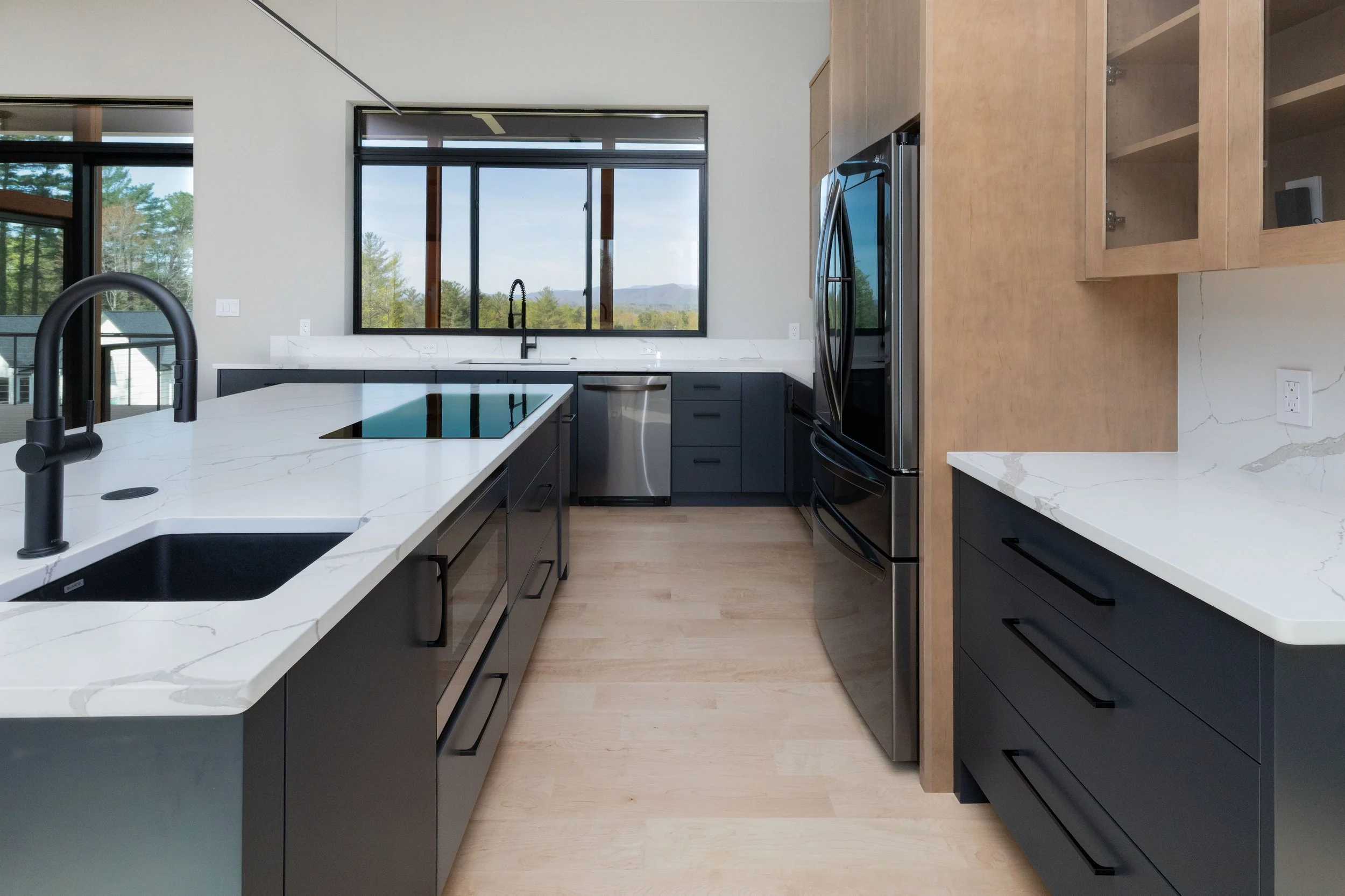 Modern kitchen with black cabinetry, white marble countertops, stainless steel appliances, and large windows showing an outdoor view.