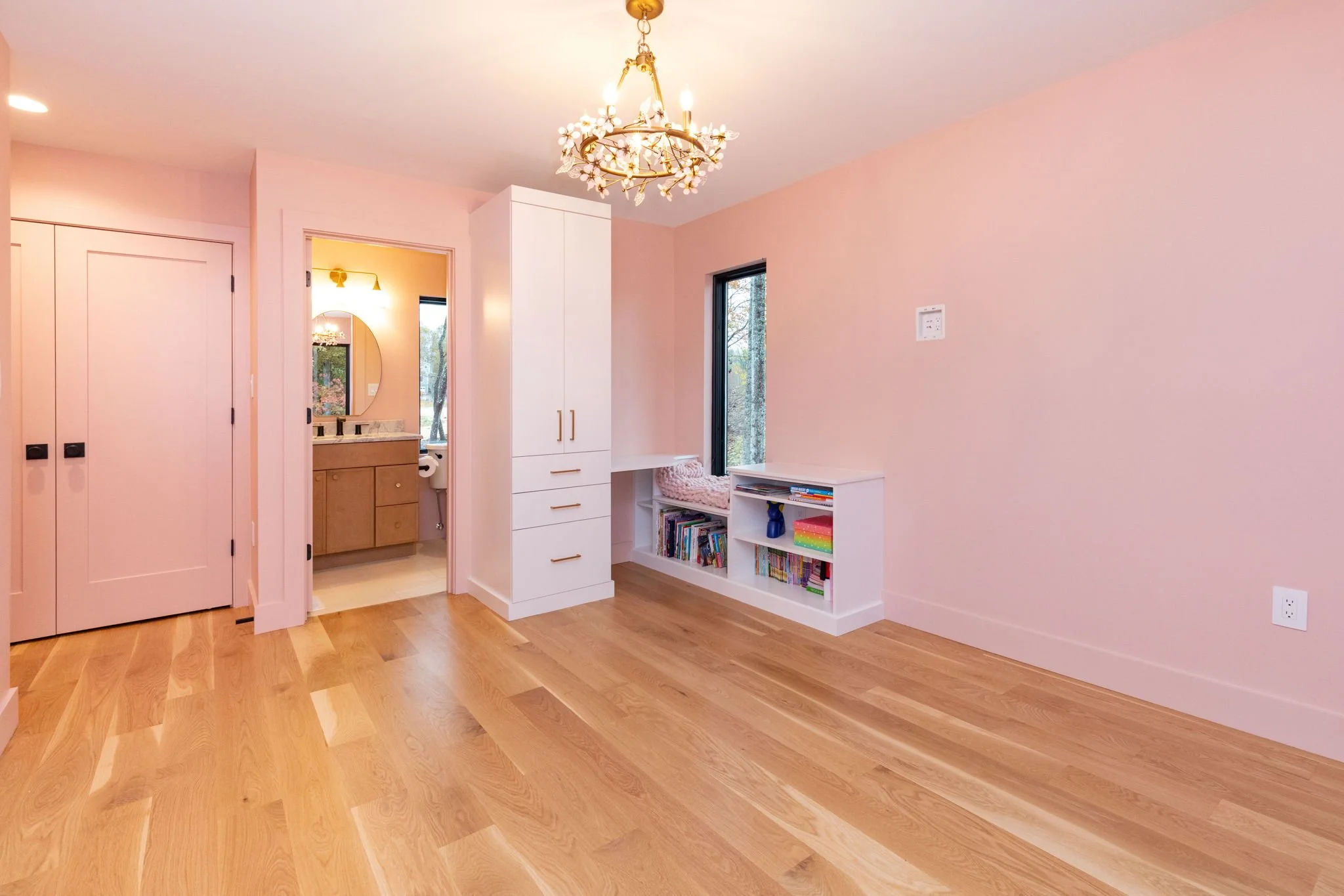 Empty pink-colored room with hardwood floors, a white built-in bookshelf, a white wardrobe, chandelier, and view into a bathroom with a vanity and mirror.