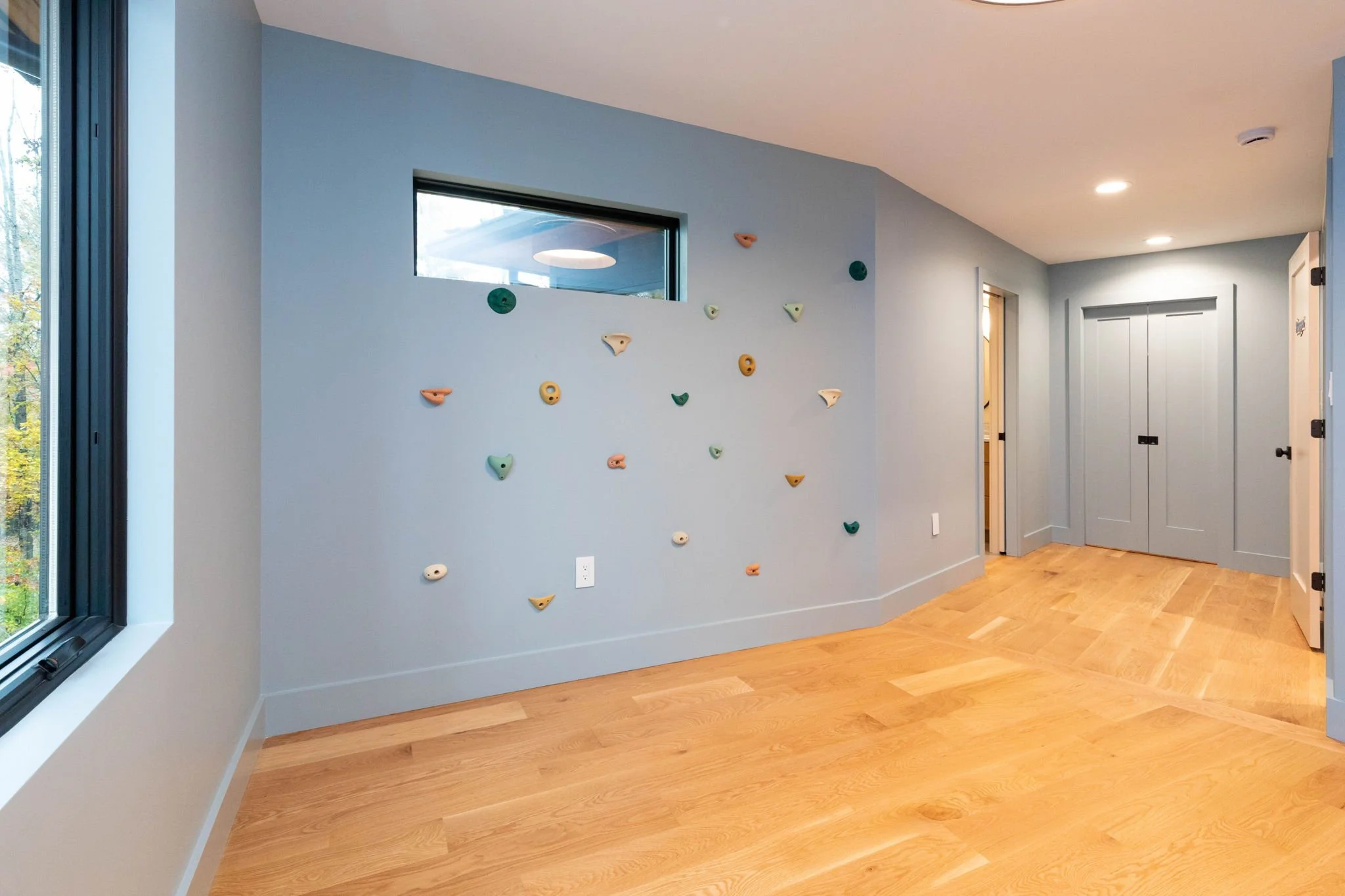 A room with light blue walls, hardwood floors, and climbing holds on one wall. There is a window with a black frame and a door with double panels in the background. Recessed ceiling lights illuminate the room.