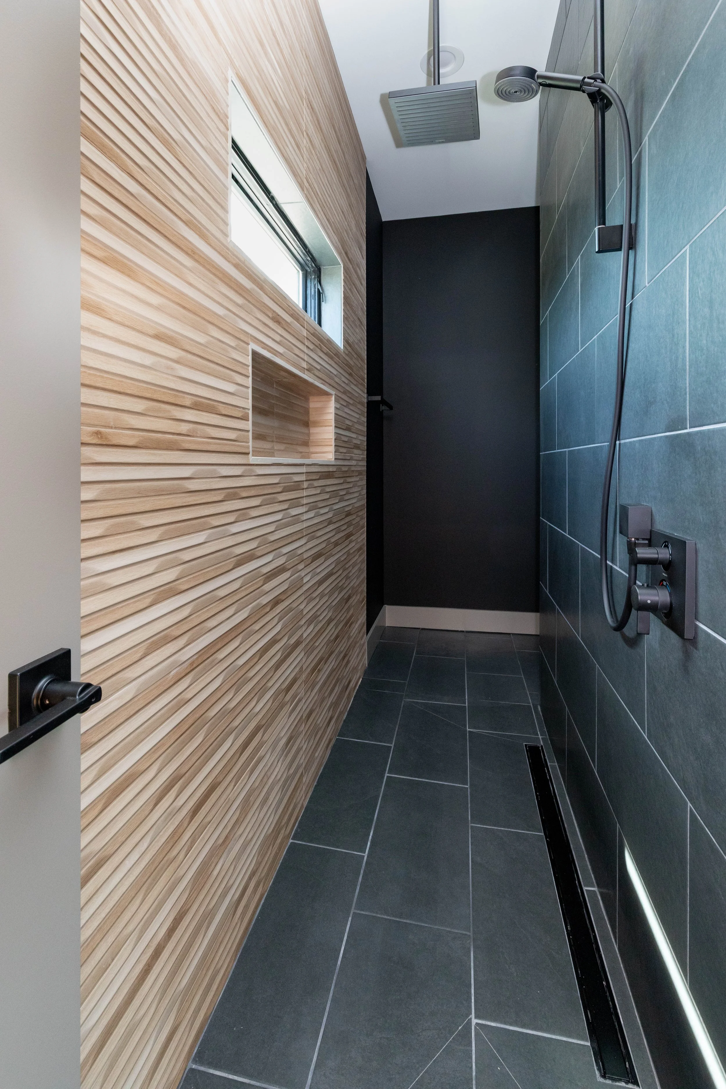 Modern shower with dark gray tiled floor and wall, black fixtures, and a wooden accent wall with a small window.