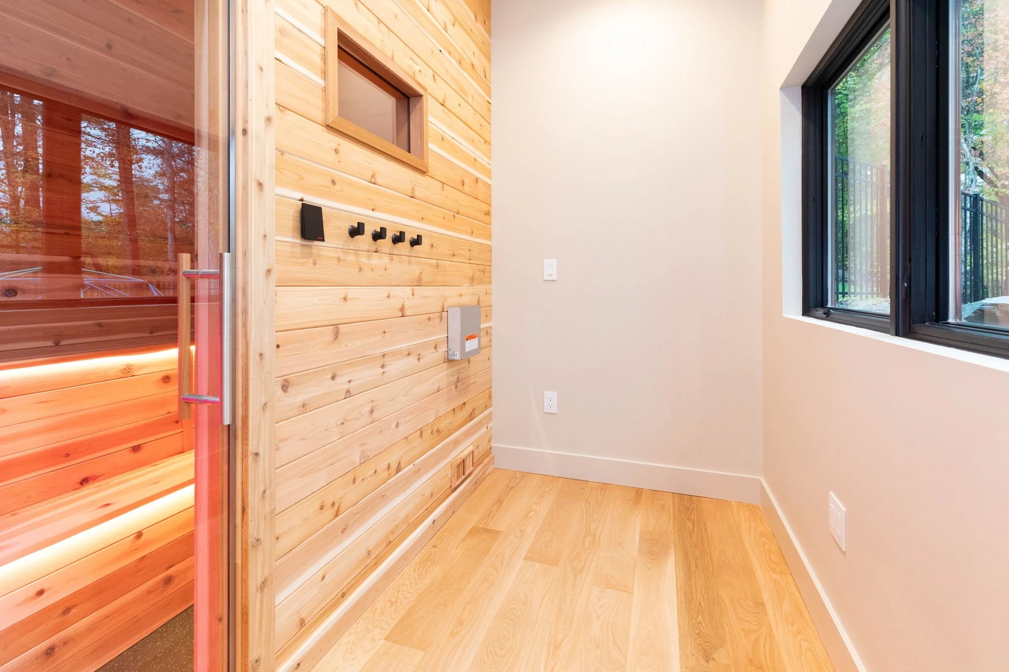 Empty room with wood-paneled wall on the left, white walls on the right, a window showing trees outside, and a wooden floor.