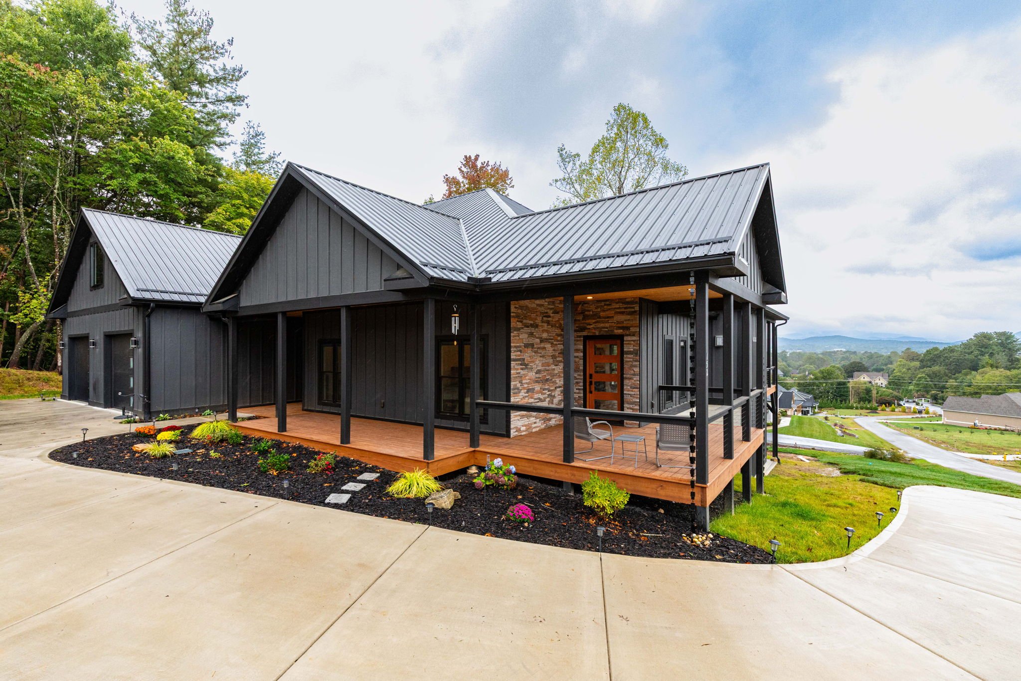 A modern black house with a metal roof, a wooden porch, and stone accent wall, surrounded by landscaped garden with colorful plants, situated on a sloped lot with concrete driveway and scenic distant view.