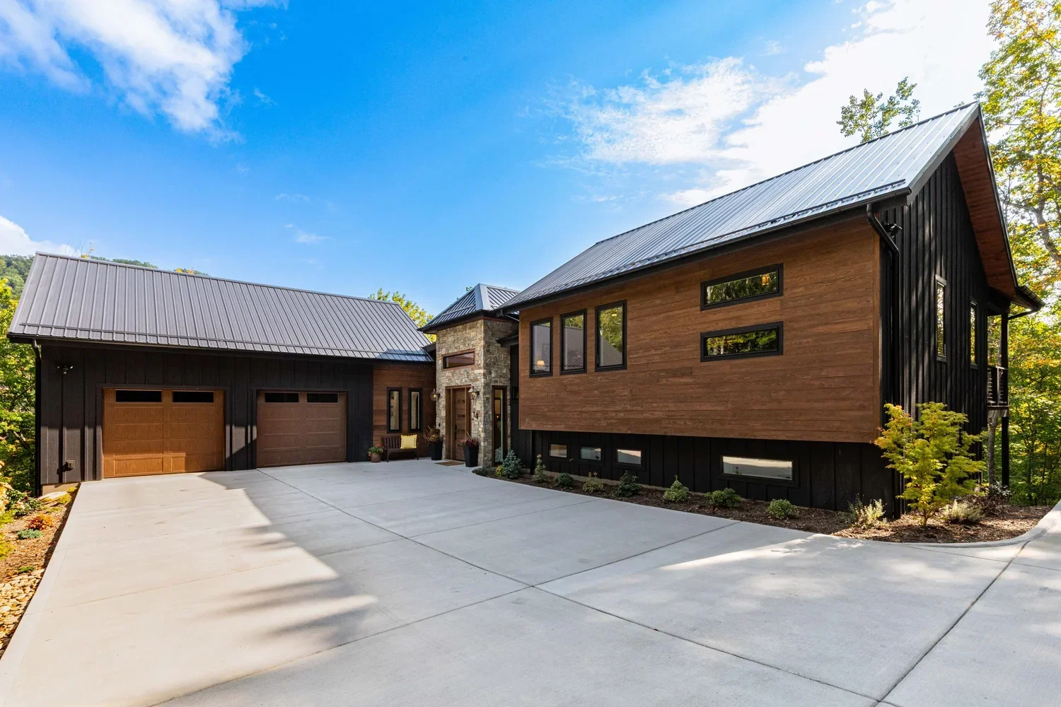 Modern house with black and wood siding, two-car garage, stone accents, well-maintained driveway, landscaped yard, and trees in the background under a blue sky with clouds.