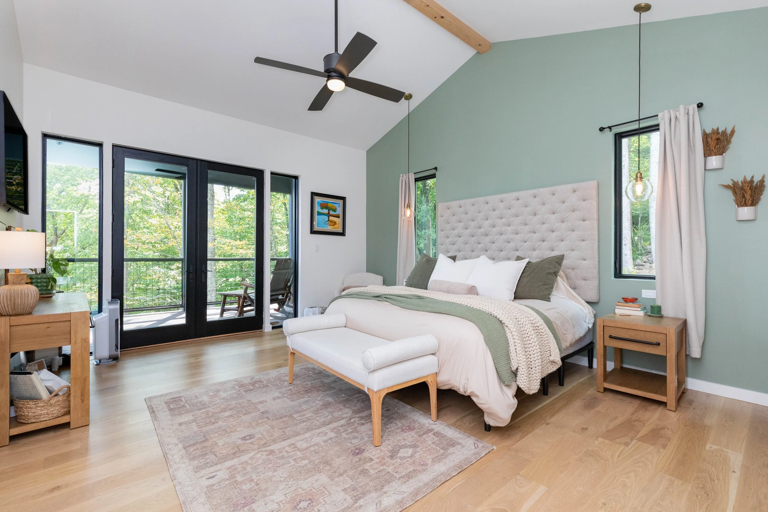Bright bedroom with a large bed, green and white decor, hardwood floors, ceiling fan, sliding glass doors leading to a balcony with outdoor seating, and windows letting in natural light.