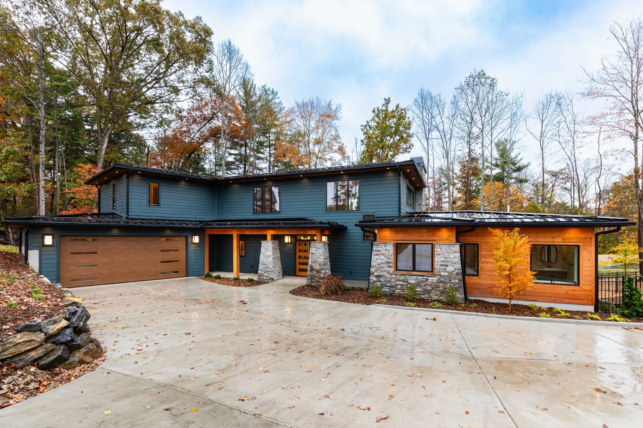 A modern two-story house with blue and wood exterior, stone accents, and a sloped driveway surrounded by autumn trees.