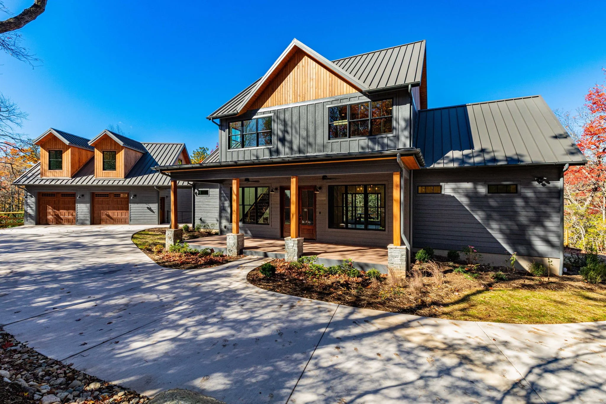 Modern two-story house with a dark gray exterior, wooden accents, and a metal roof, surrounded by a landscaped yard with a curved concrete driveway and trees.